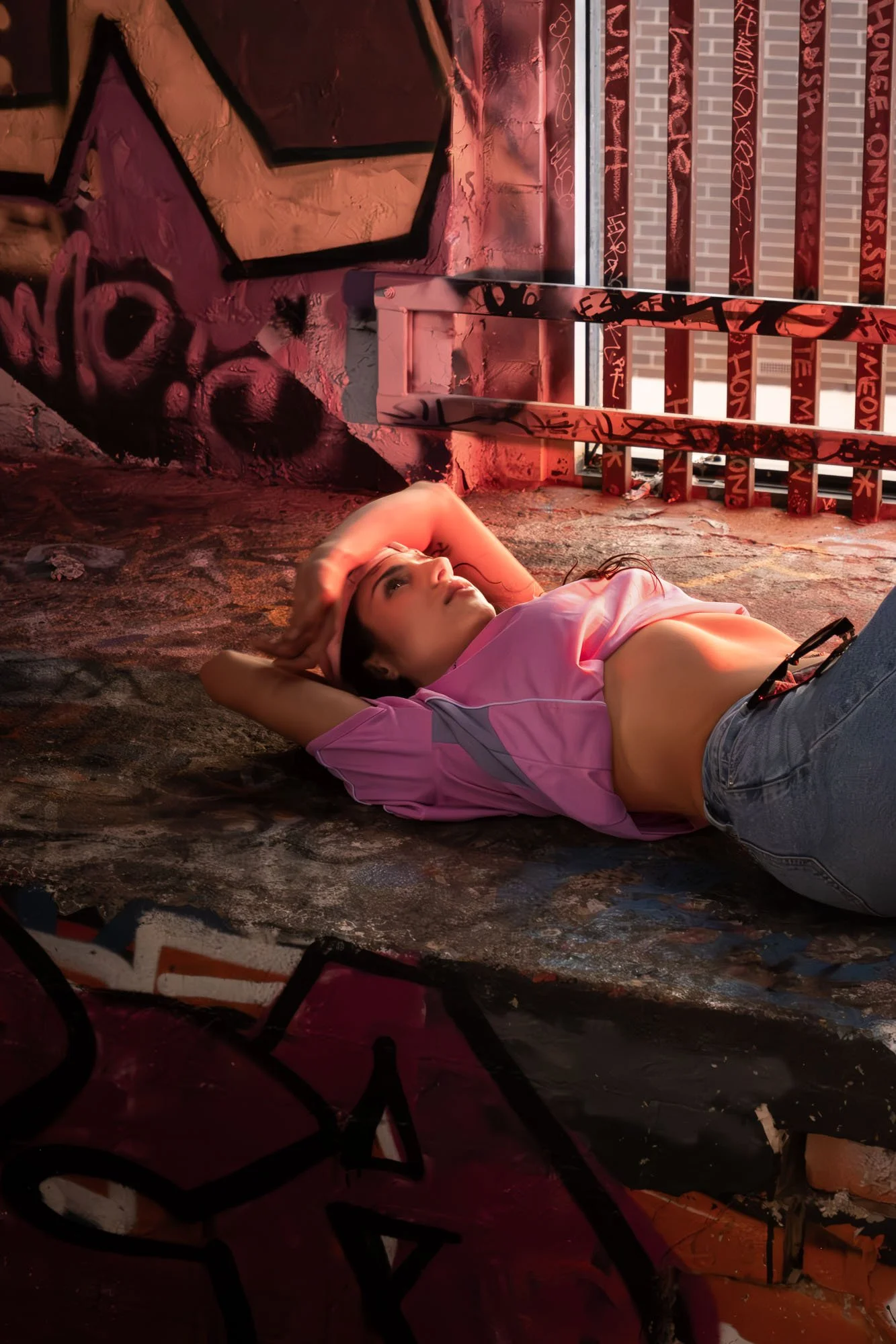 A young woman lying on the ground in an alleyway with graffiti-covered walls and a metal railing behind her, bathed in pinkish-red lighting.
