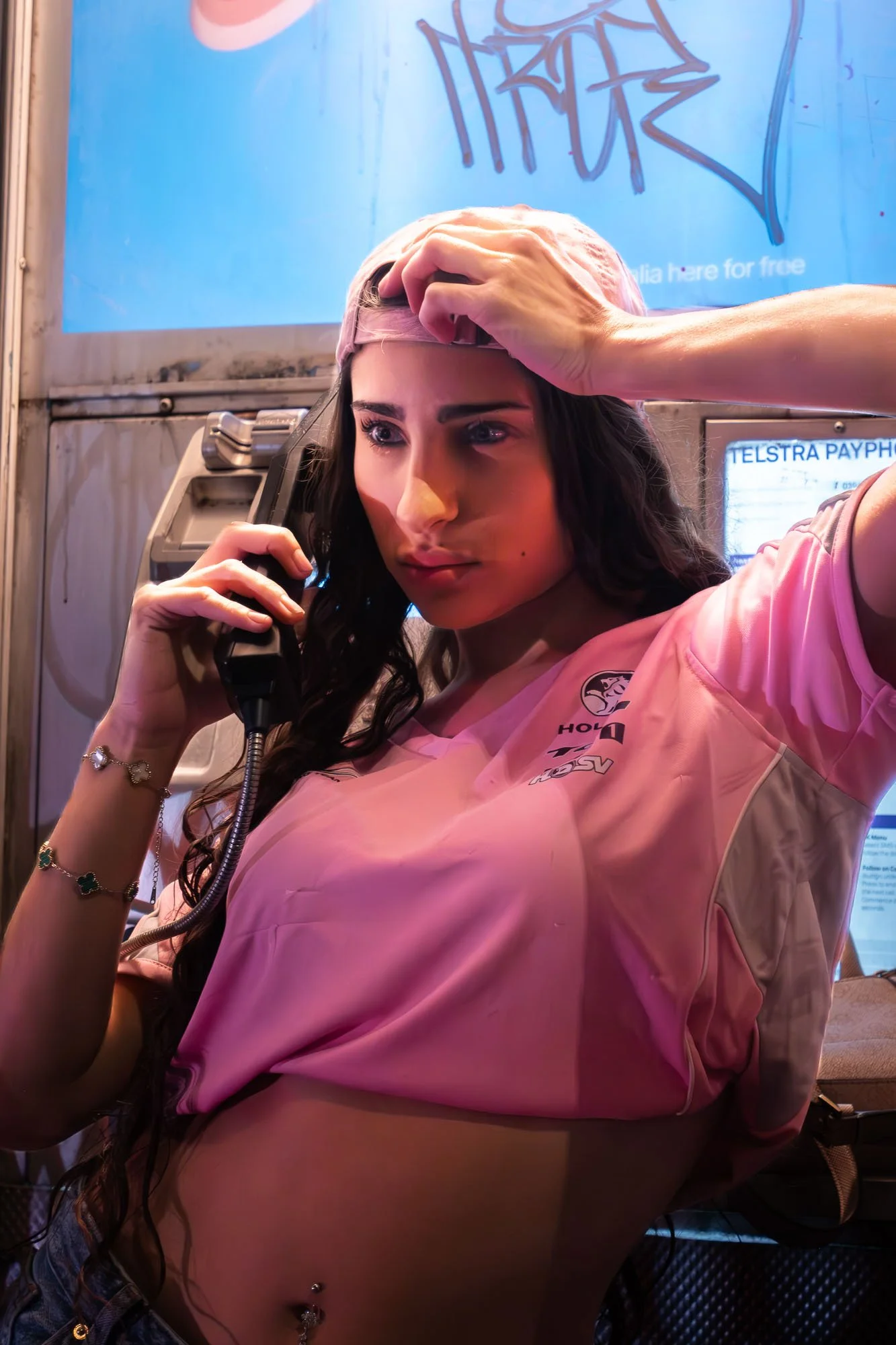 A woman with dark hair and fair skin talking on a landline telephone, wearing a pink sports jersey and a pink cap, with her hand on her head. A tattoo and a navel piercing are visible, and she wears multiple bracelets on her wrist. She is in a settin