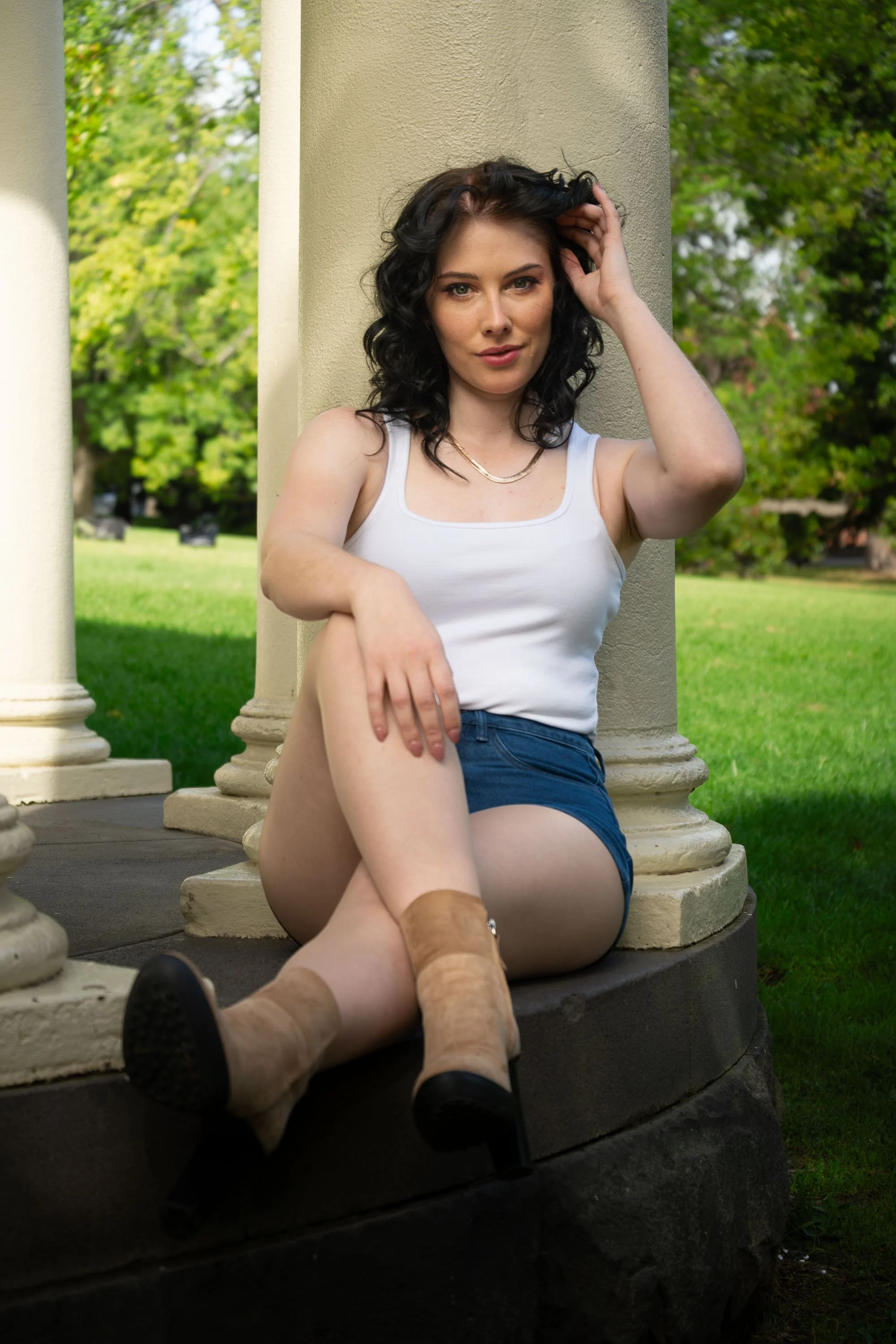 A woman with black curly hair, wearing a white tank top and blue shorts, sitting on the ground leaning against a beige column, outdoors in a park with green trees and grass in the background, with her legs crossed and one hand touching her hair.