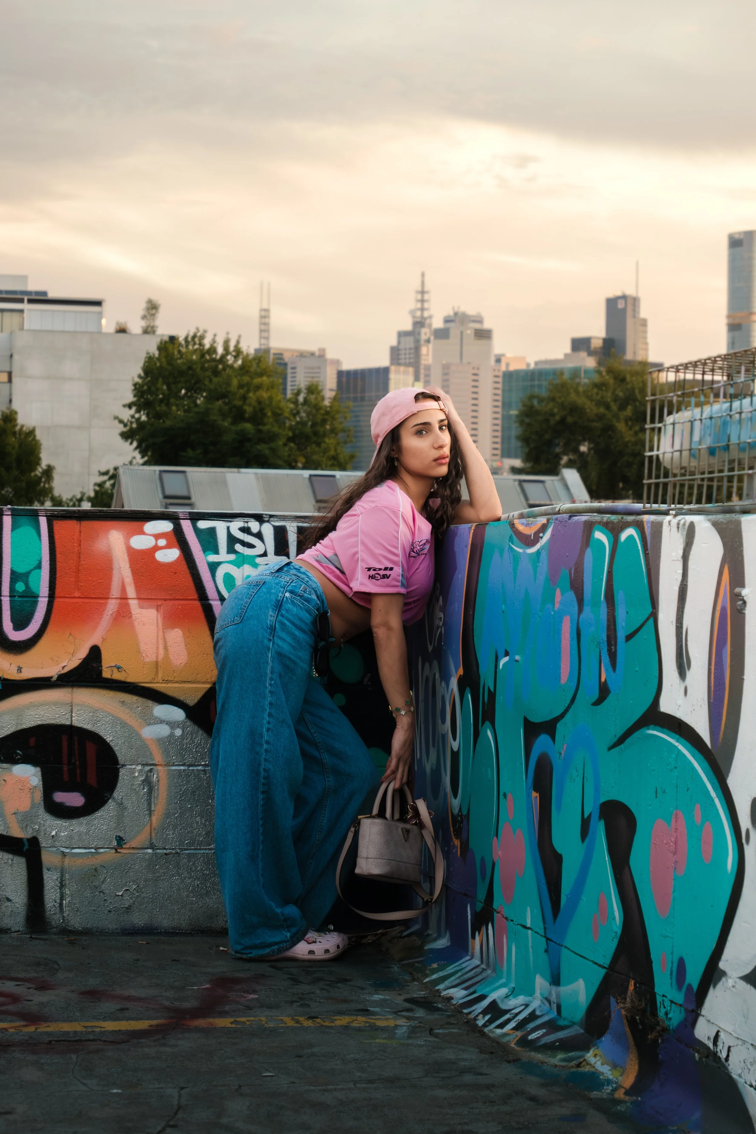 Young woman wearing a pink baseball cap and pink crop top posing against a graffiti-covered wall in an urban rooftop setting with city skyscrapers in the background.