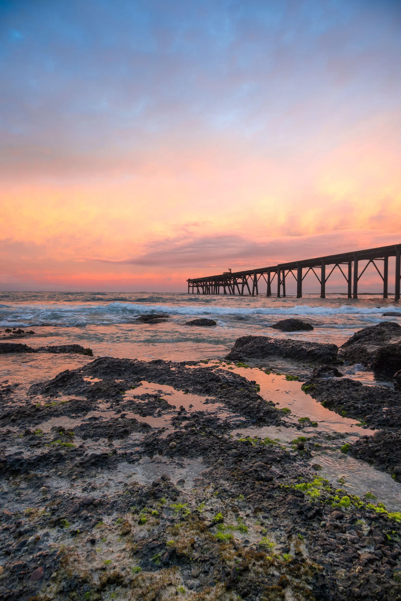 Sunset over the ocean with a pier extending into the water and rocky shoreline in the foreground.