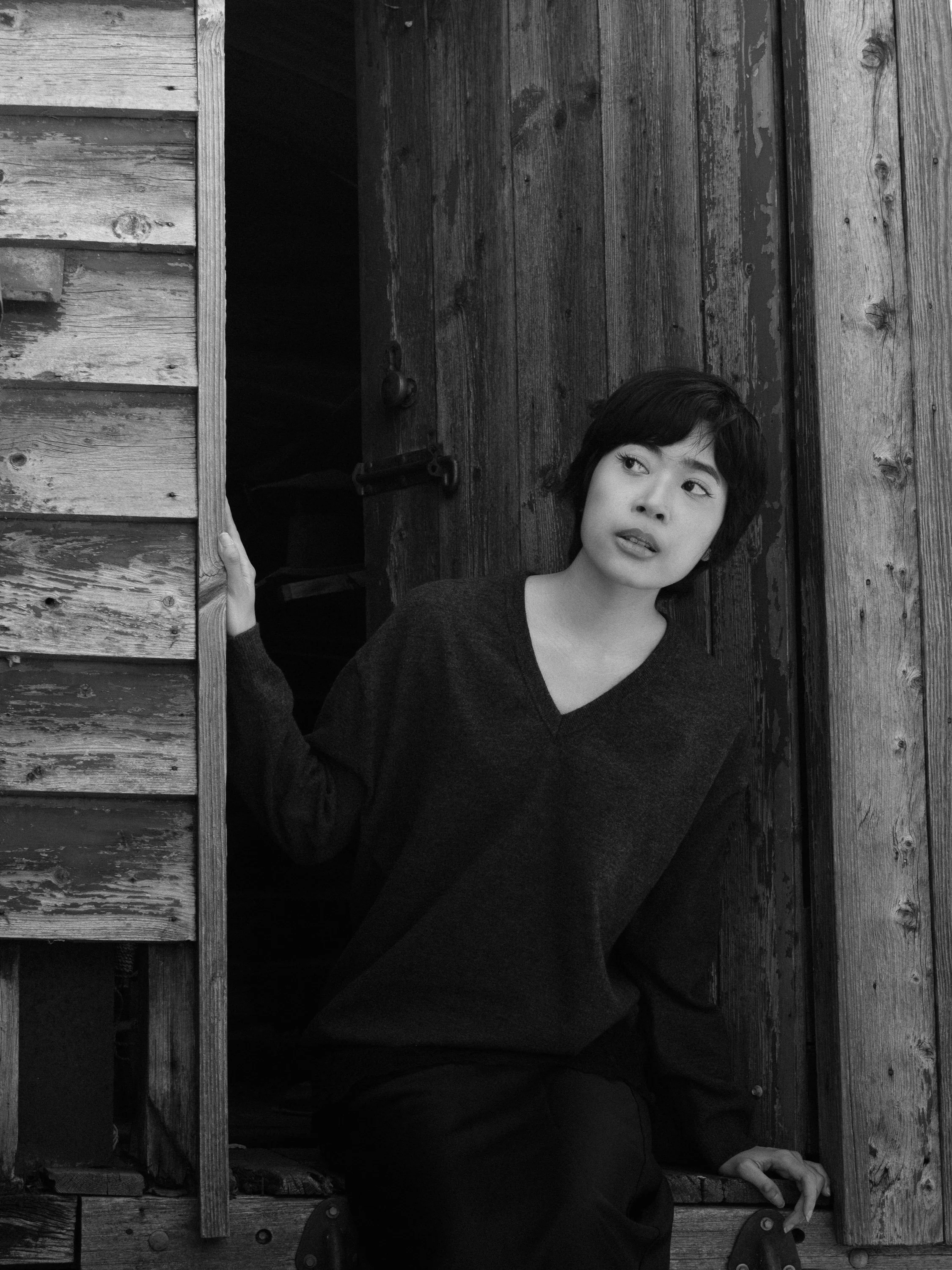 A woman with short dark hair and a dark top is looking to her right while peeking out from a wooden structure, with her hand on the edge of the structure.