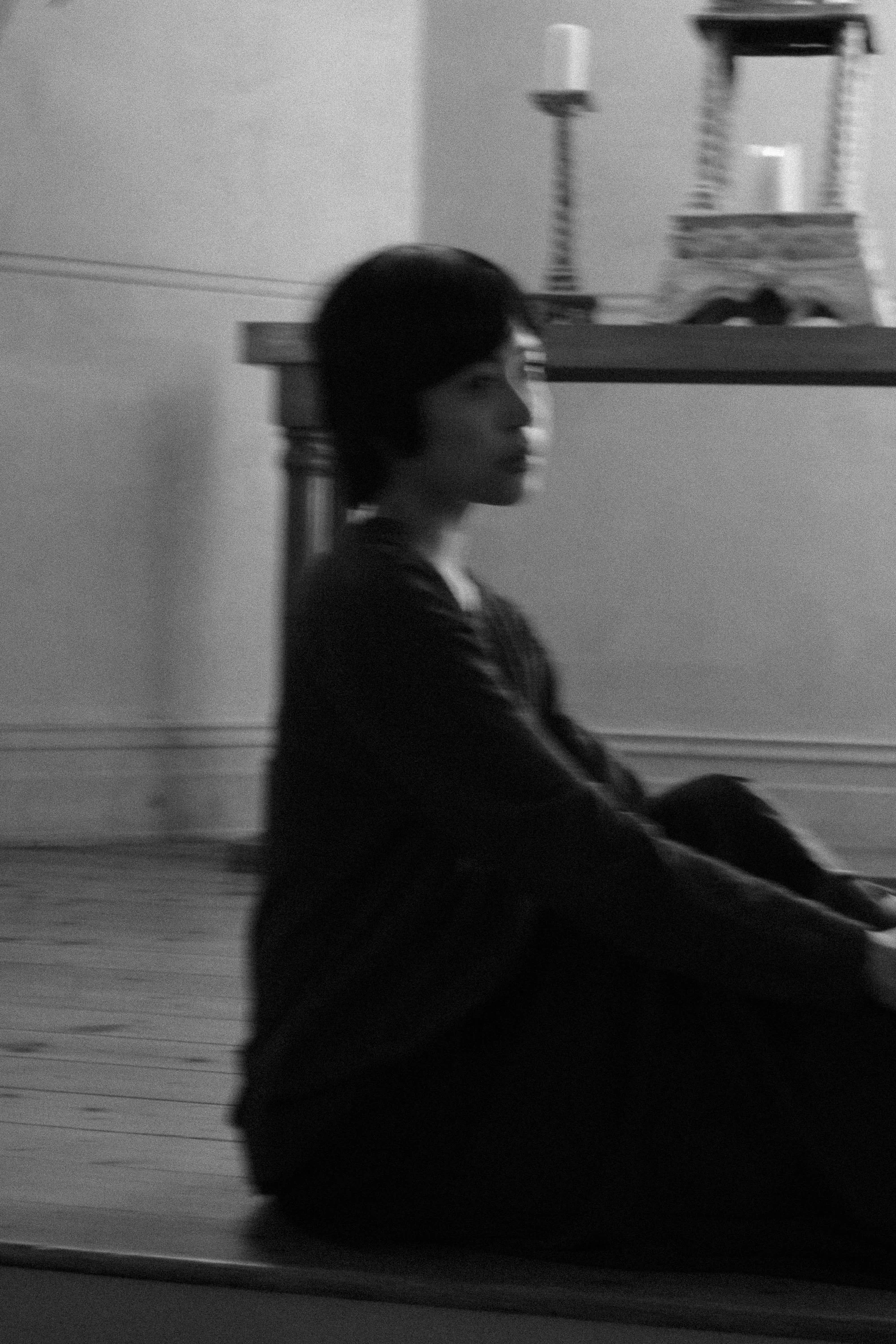 A person with black hair sitting on the ground indoors, leaning against a wall with shelves and objects. The image is in black and white.