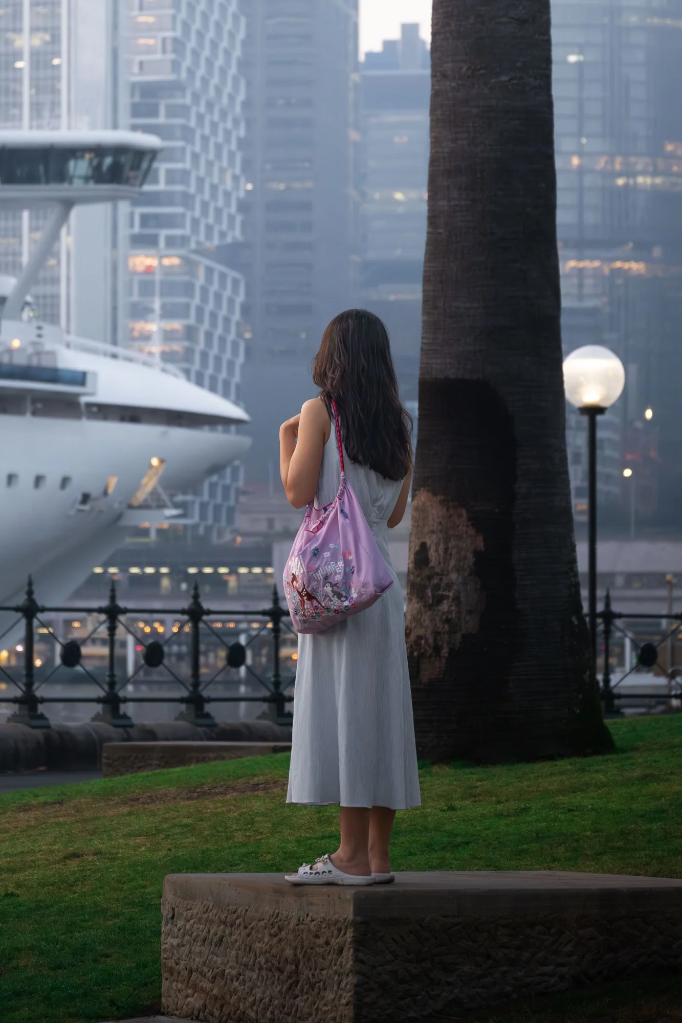 A woman stands on a raised stone platform in a city park, facing away from the camera. She wears a long white dress, white sneakers, and carries a pink tote bag with cartoon characters. The background features modern high-rise buildings, a large whit