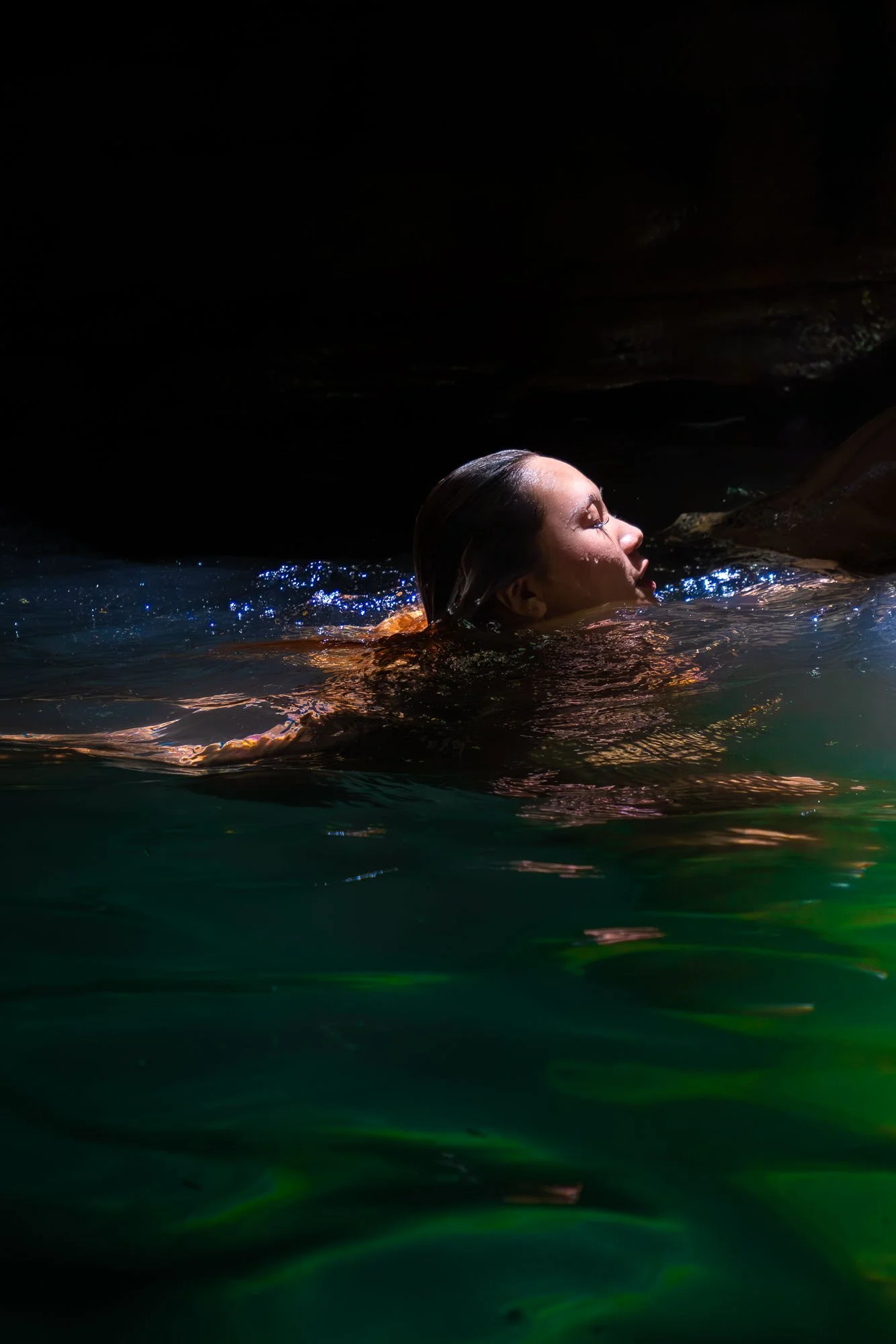 A woman with wet hair swimming in dark water, illuminated by light reflecting off the water.
