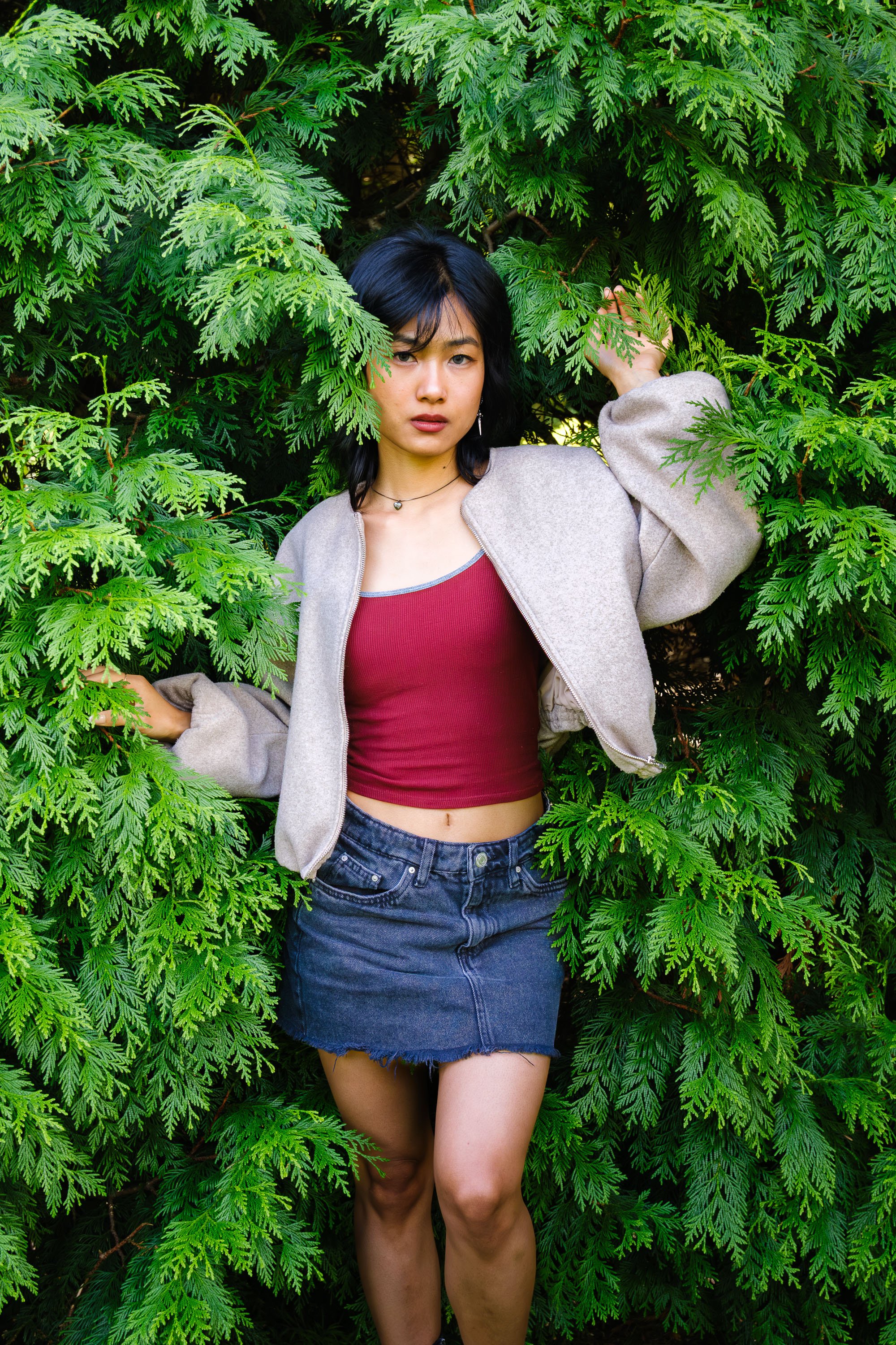 A woman with black hair and light skin standing among green foliage, wearing a red crop top, a beige jacket, and a denim skirt.