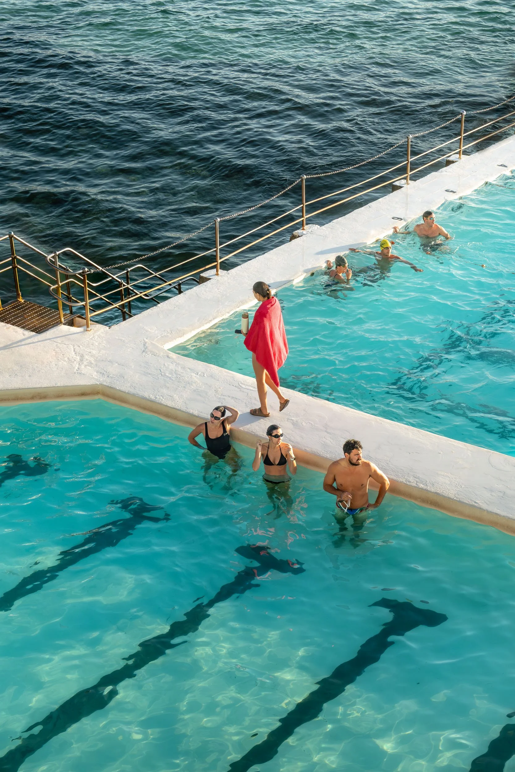 People swimming in a pool near the ocean with a person standing on the pool edge holding a water bottle.