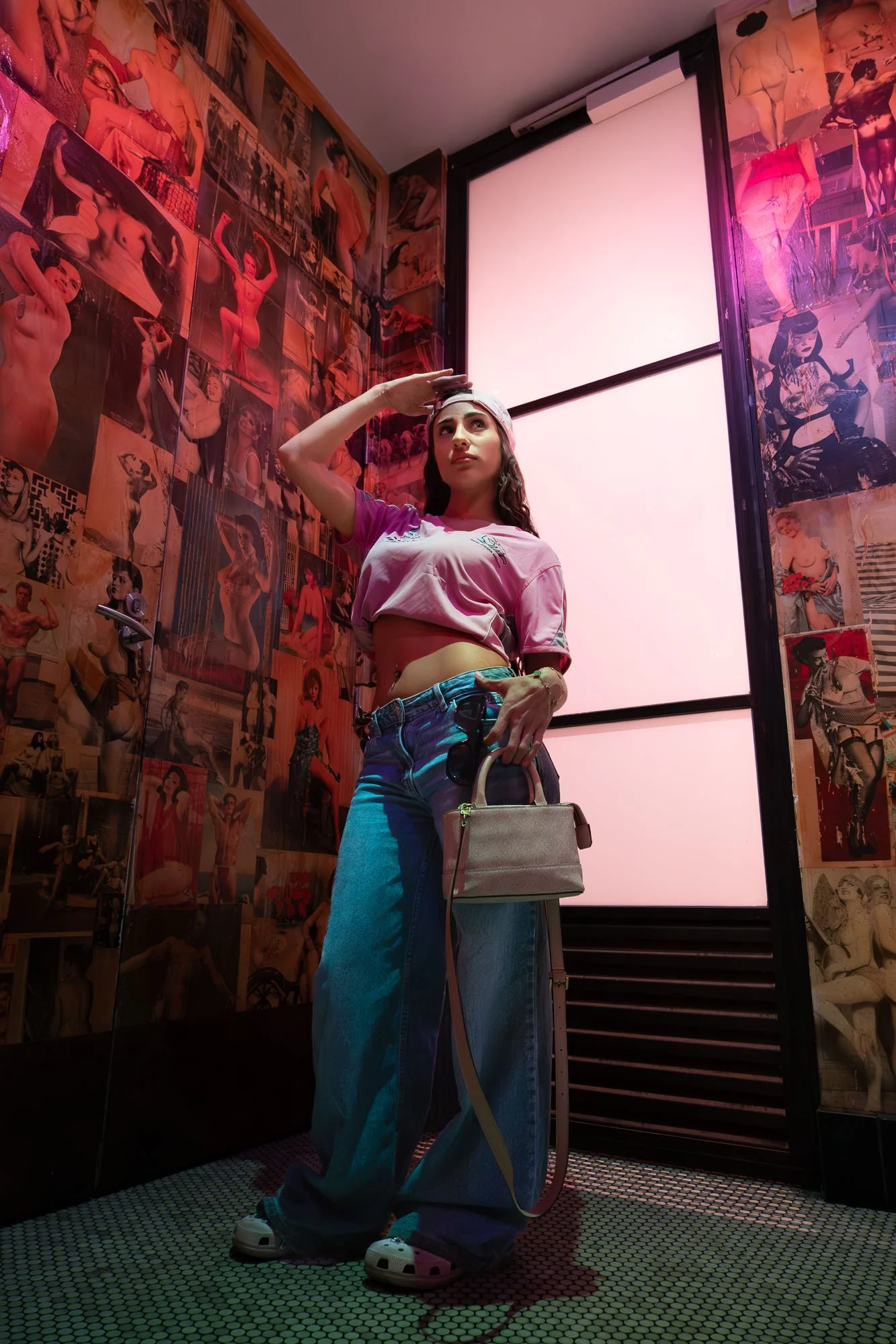 A young woman with long dark hair wearing a pink cropped t-shirt, blue baggy jeans, and white shoes, posing in front of a wall covered with various pin-up style photographs. She is tilting her head slightly upwards, holding her right hand to her fore