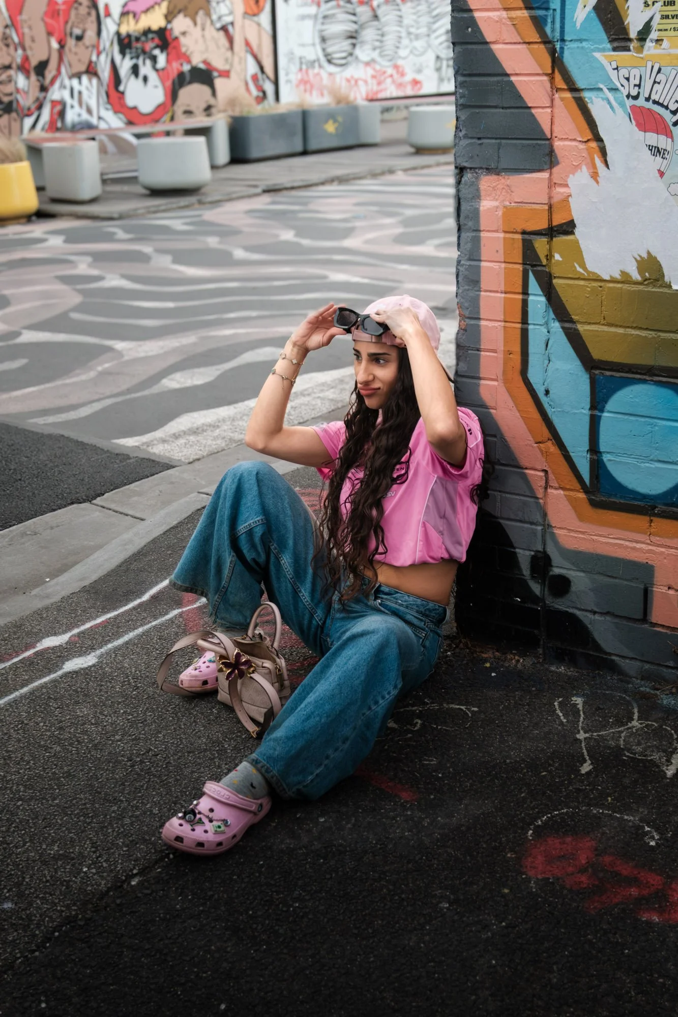 A young woman with long, curly dark hair, wearing a pink cap and pink t-shirt, sits on the ground leaning against a colorful graffiti-covered brick wall in an urban area. She is adjusting her sunglasses, with pink Crocs, blue jeans, and a small handbag placed nearby, and white sneakers on her feet with socks.