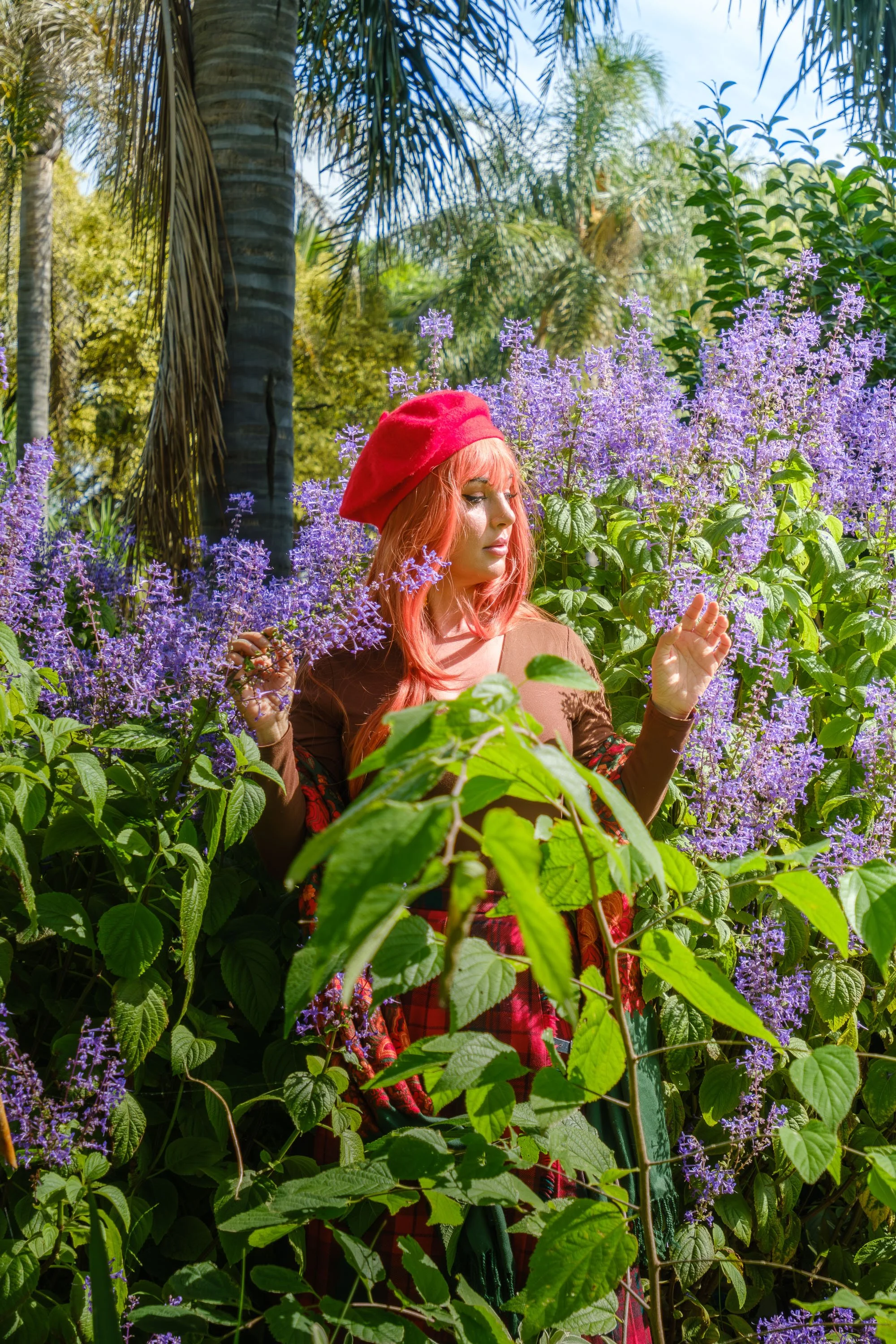 A woman with red hair wearing a red beret and colorful clothing, standing among purple flowering plants in a lush garden with trees and blue sky in the background.