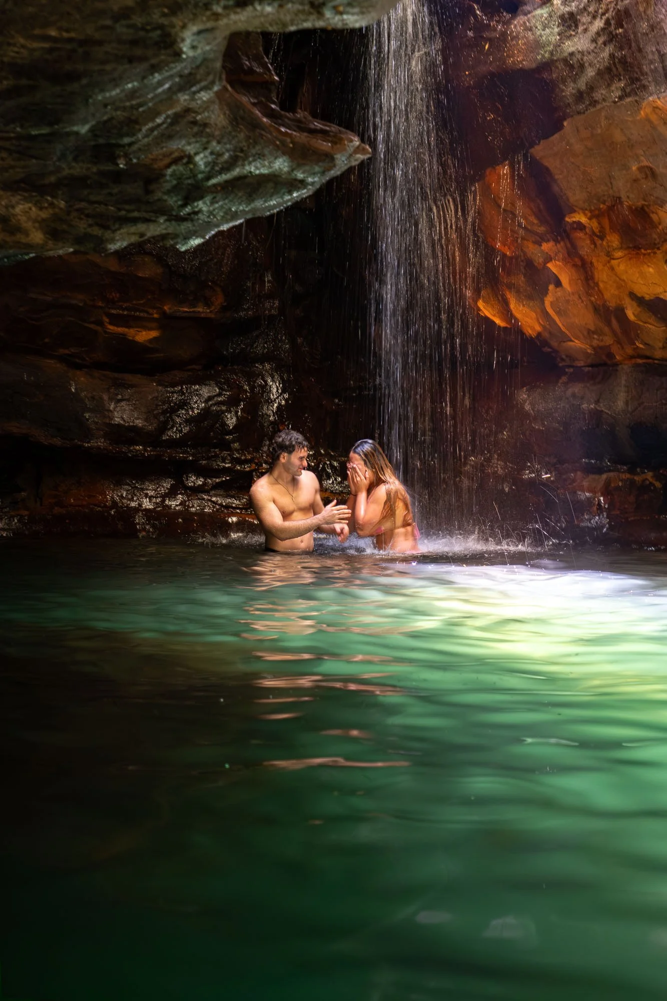 A young man and woman are standing in a natural pool under a waterfall, with rocky cliffs around them. The woman covers her face while the man presents something in his hands.