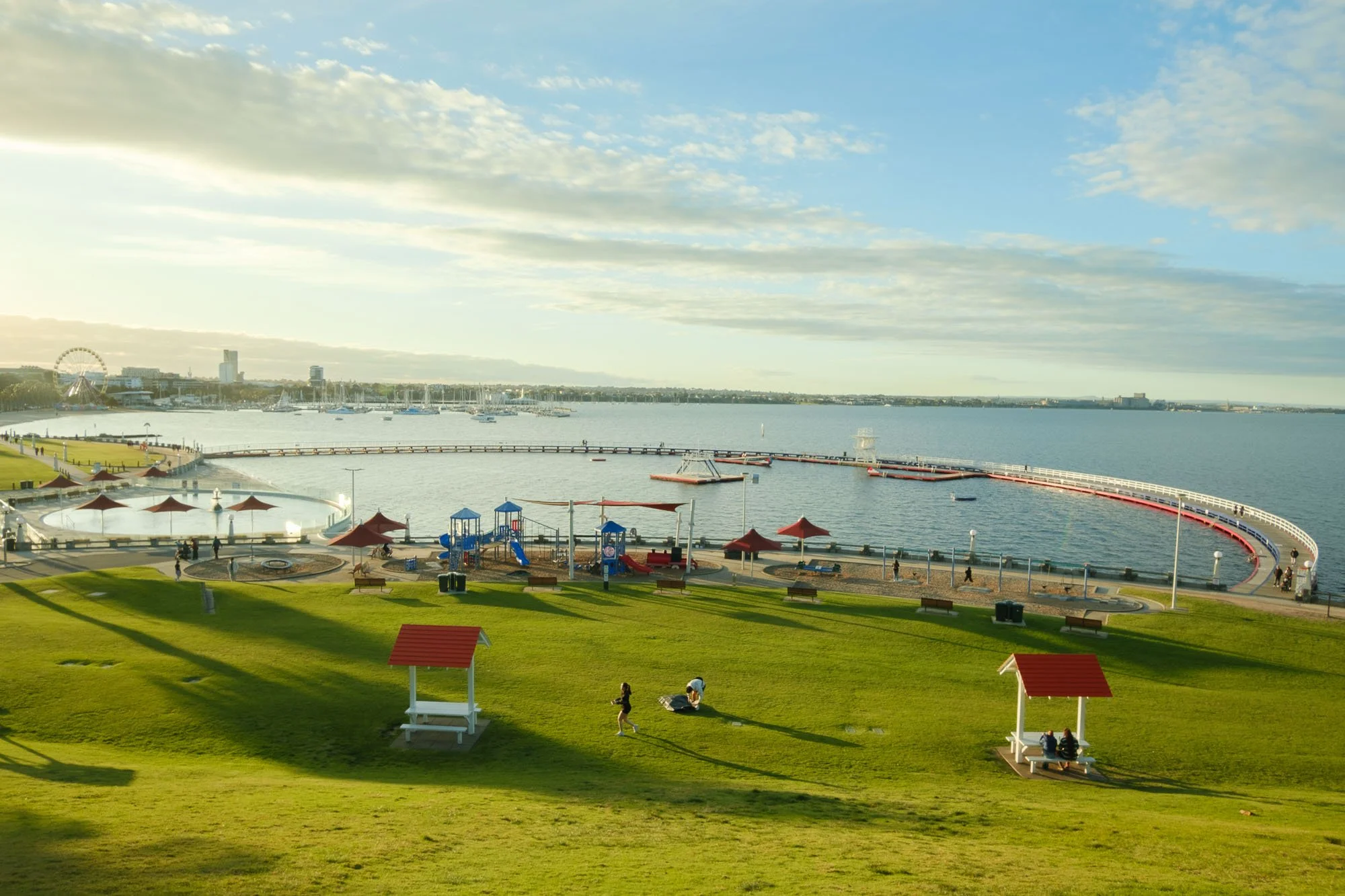 A seaside park with green grass, benches, small red-roofed structures, and a playground area. Behind the park, there is a shoreline with a harbor featuring boats, a ferris wheel, and high-rise buildings in the distance. The sky is partly cloudy with sunlight illuminating the scene.