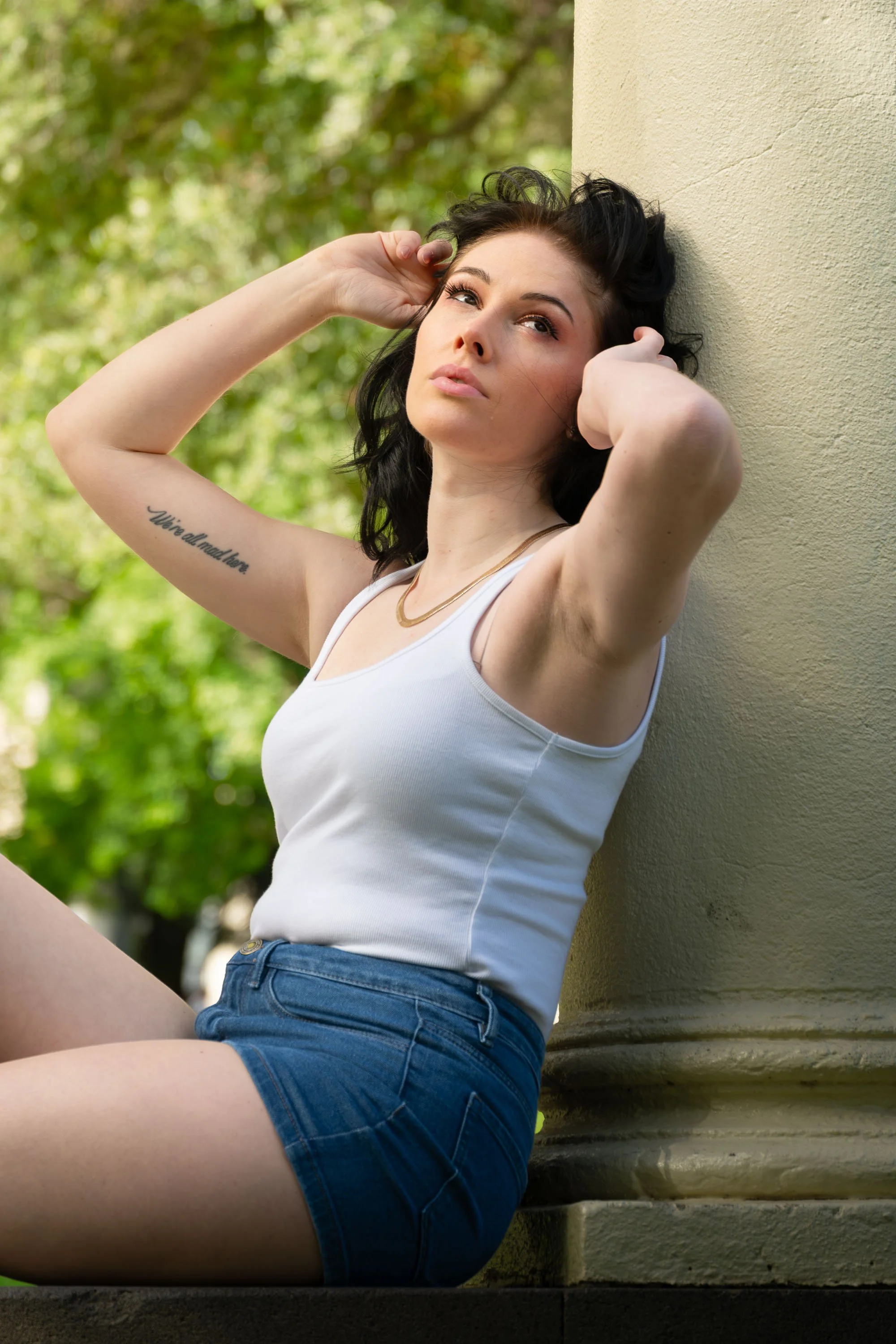 A woman with black hair and a tattoo on her arm sitting against a beige column outdoors, looking thoughtfully into the distance, wearing a white tank top and denim shorts.