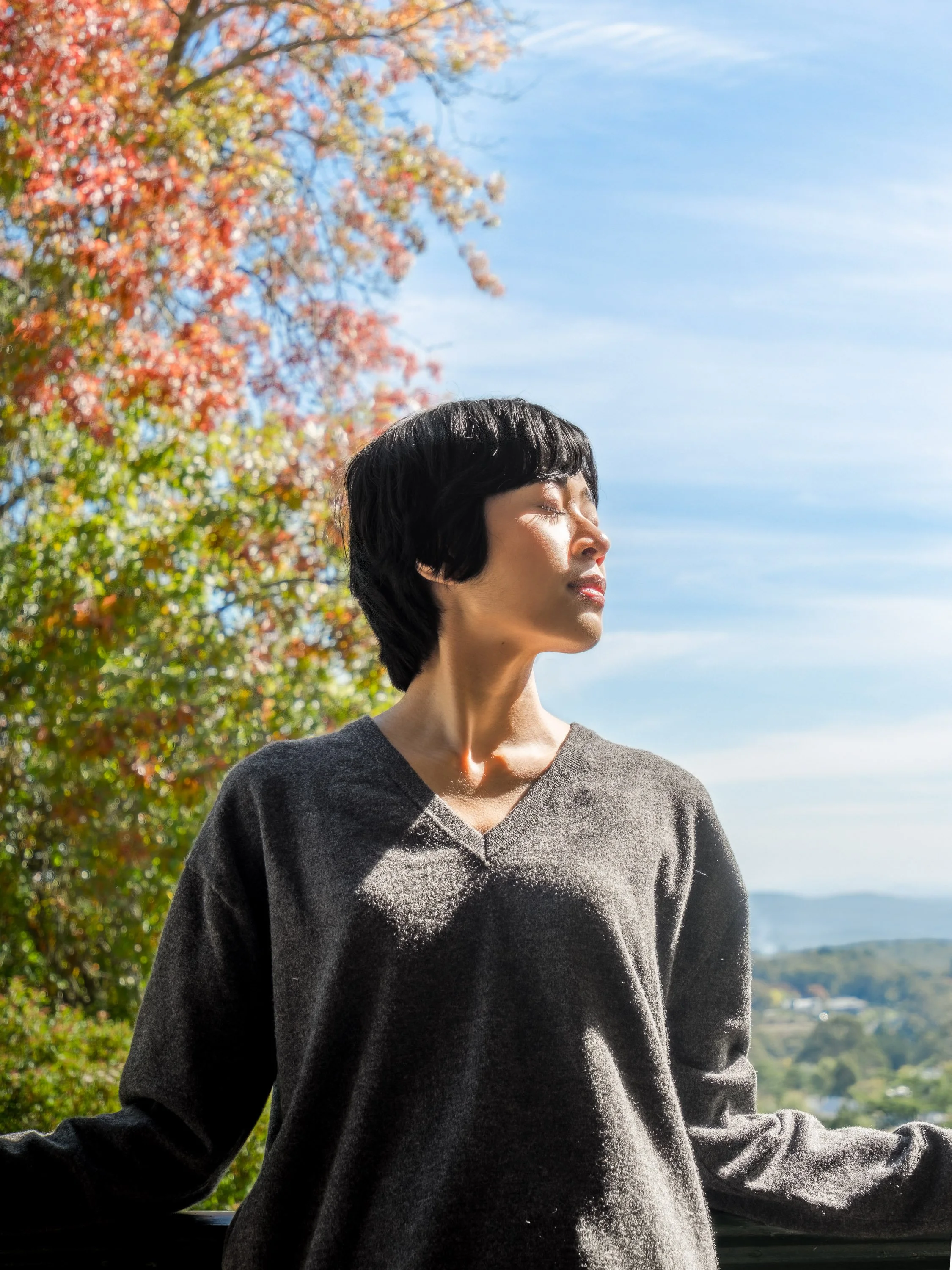 A woman with short black hair is standing outdoors with eyes closed, facing the sun. She is wearing a dark gray V-neck sweater. Behind her, there are trees with colorful fall foliage, partly blurred, and a blue sky with some clouds. In the background