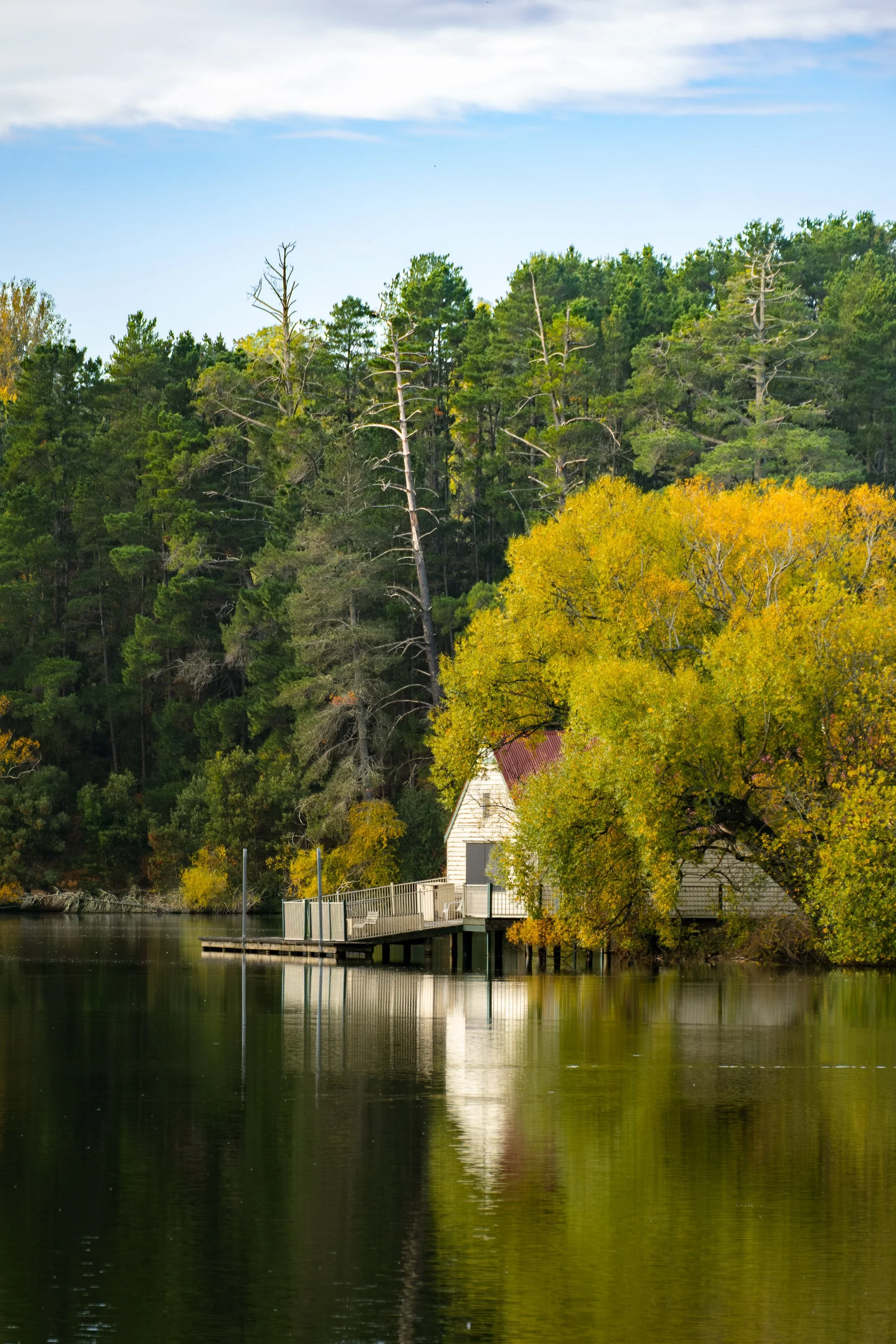 A lakeside scene with a small house, surrounded by trees with autumn foliage, and a forested hill in the background under a partly cloudy sky.