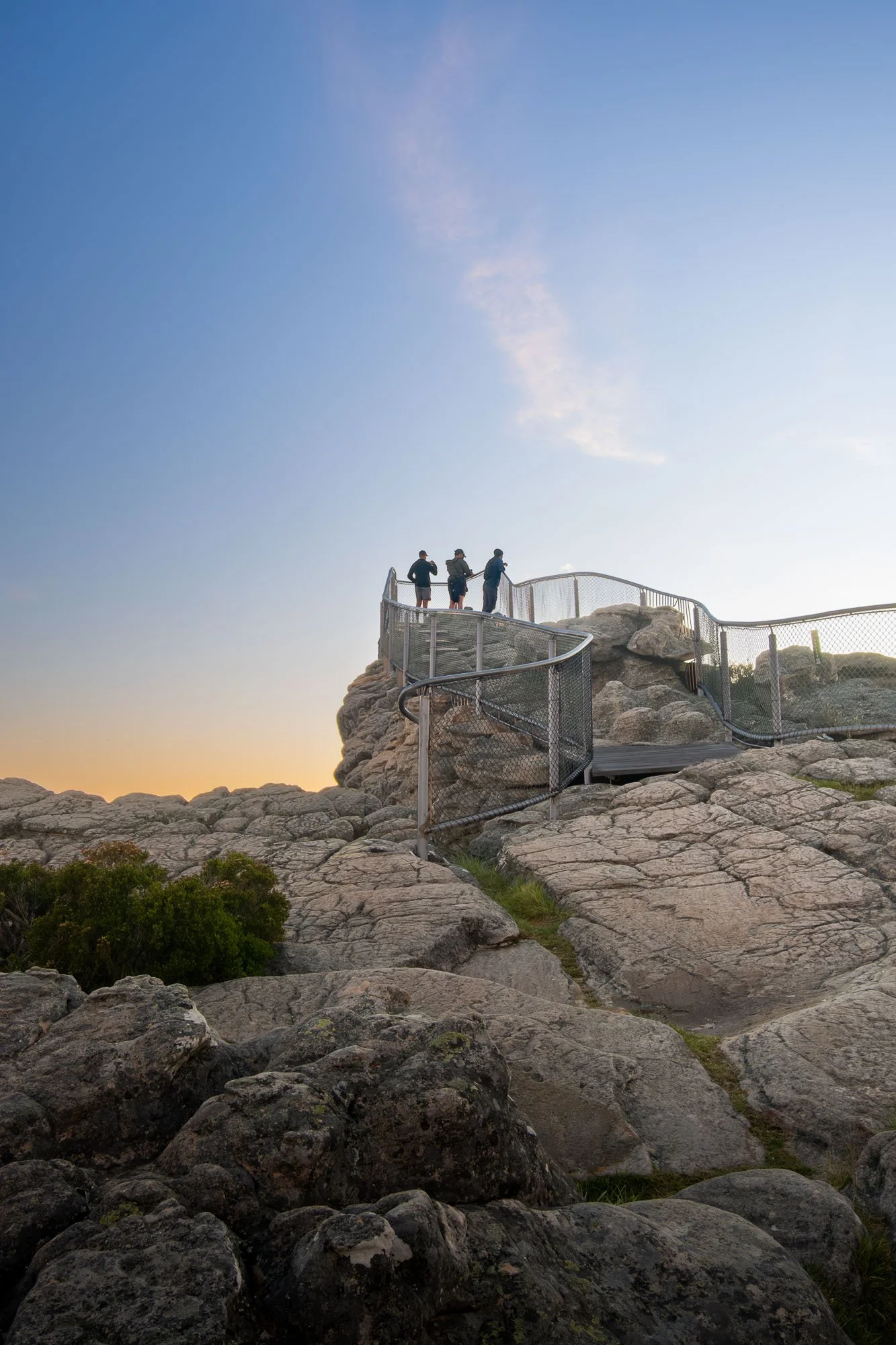 Four people standing on a rocky cliff with metal railing at sunset, overlooking the landscape.