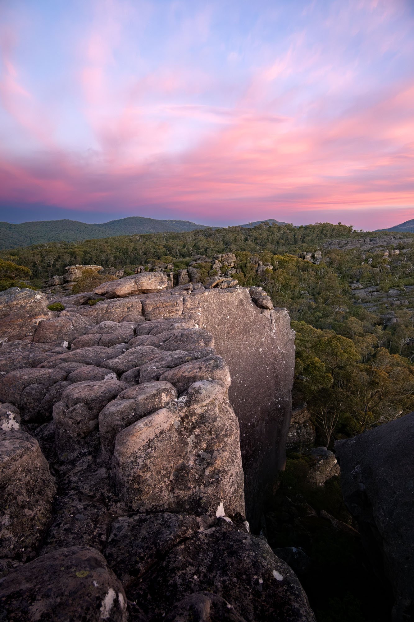 A landscape view of rocky terrain and trees with distant hills under a pink and blue sky at sunset.