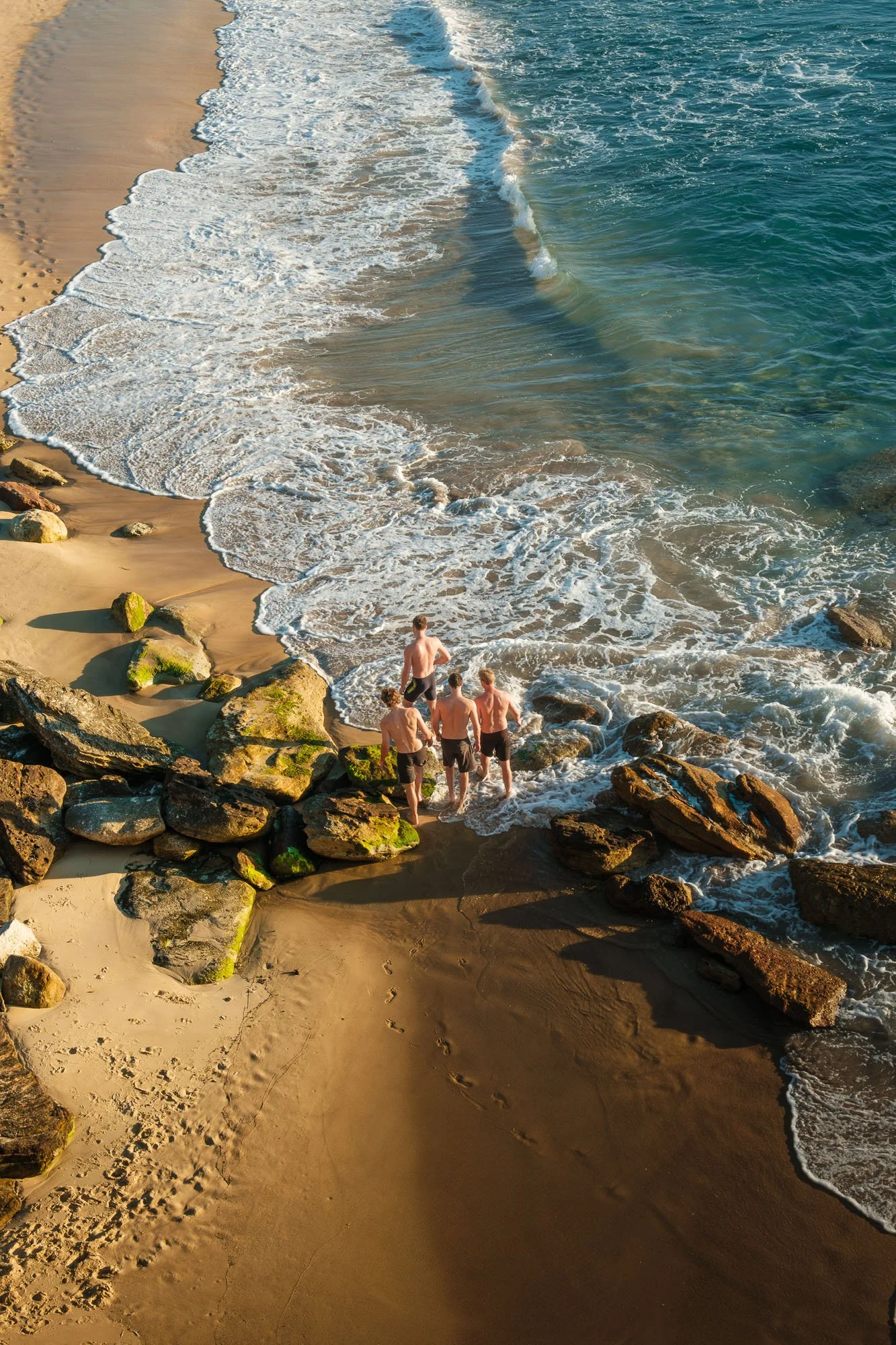 Four shirtless men walking on rocks and sandy beach near the ocean, with waves crashing onto the shore during daylight.