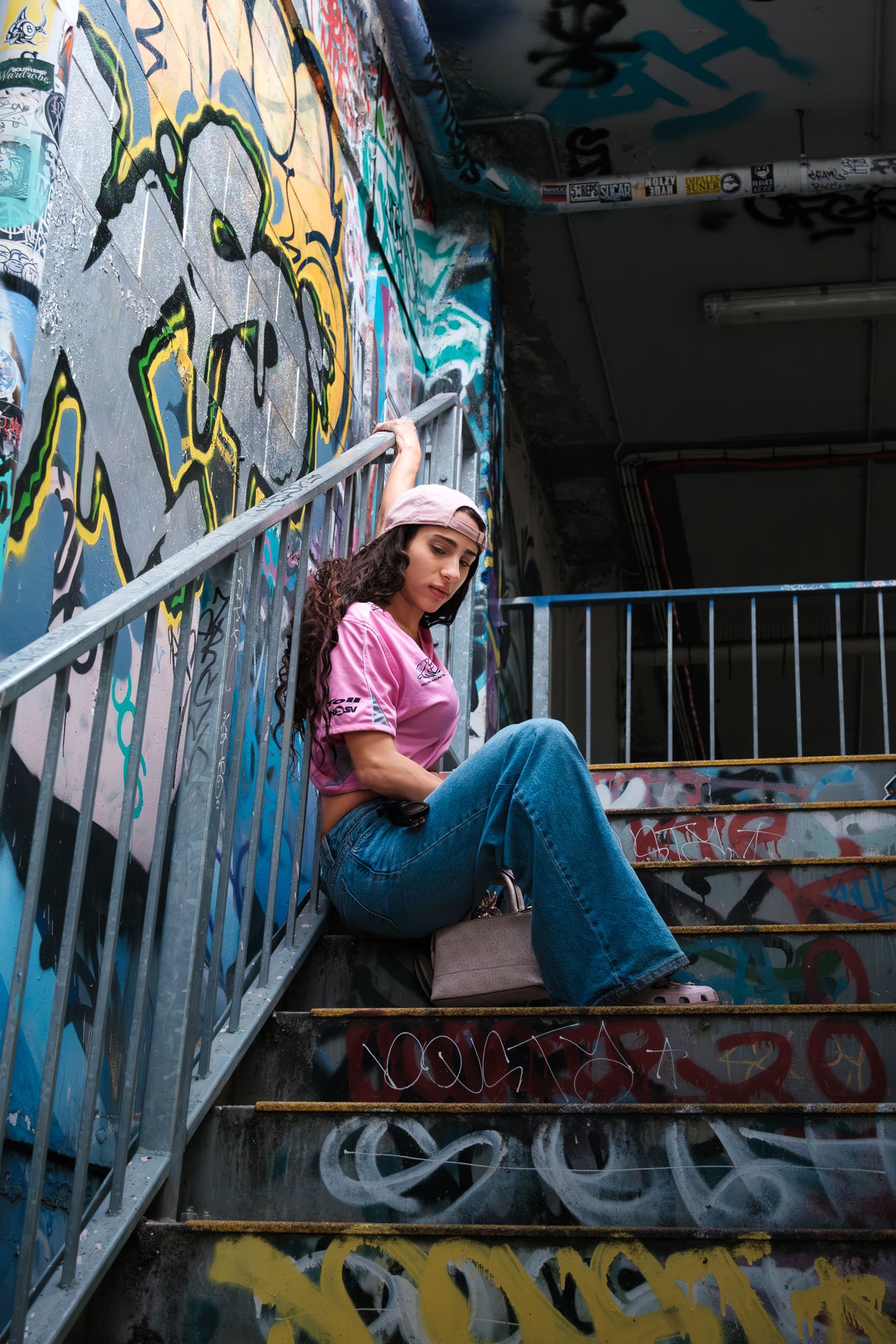 A young woman with long curly hair, wearing a pink cap, pink T-shirt, and blue jeans, is sitting on graffiti-covered stairs with a metal railing. She looks down thoughtfully while holding onto the railing with her right hand, in an urban setting.