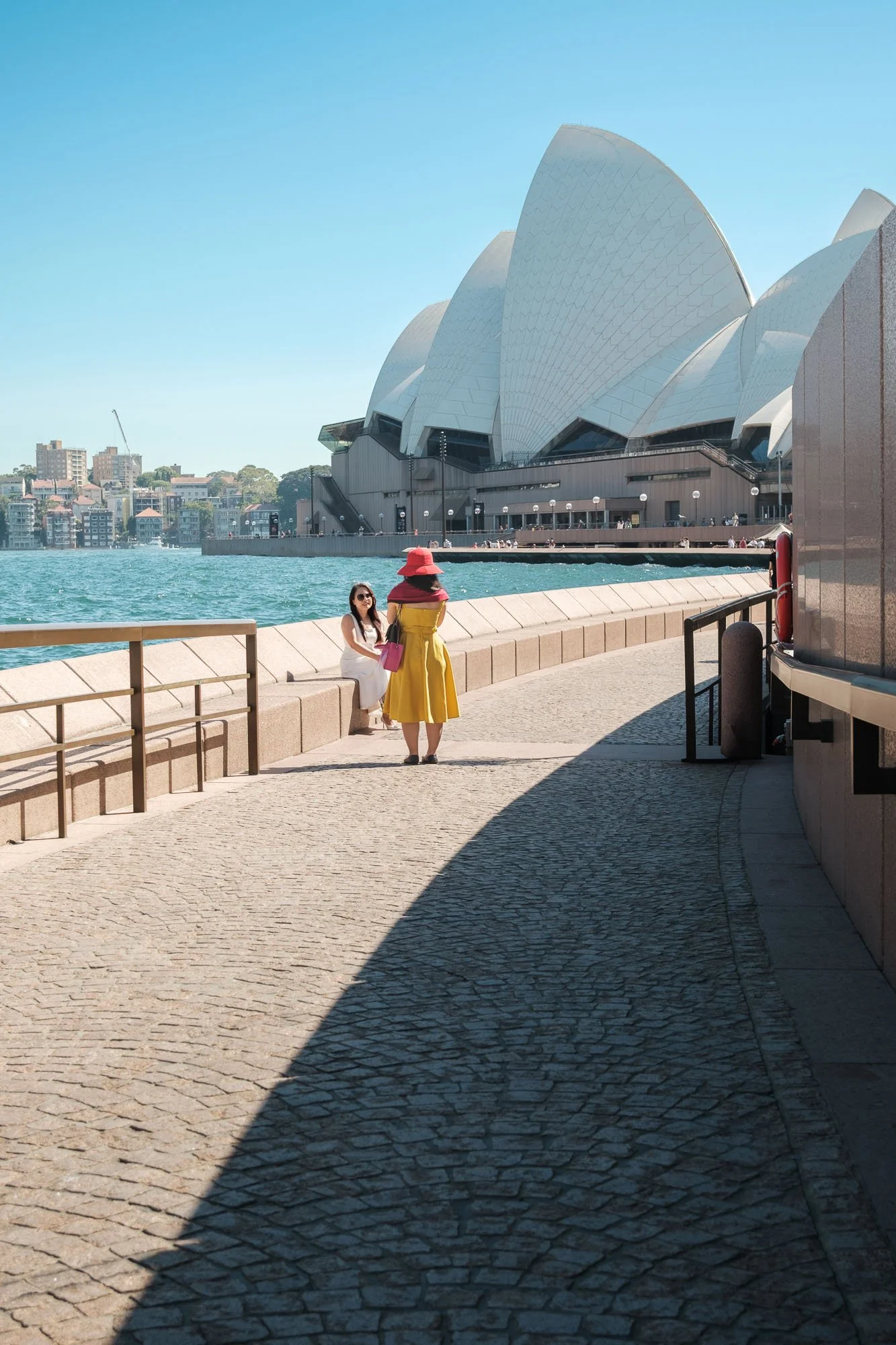 The Sydney Opera House with two women in the foreground, one sitting and the other standing, near the water on a sunny day.