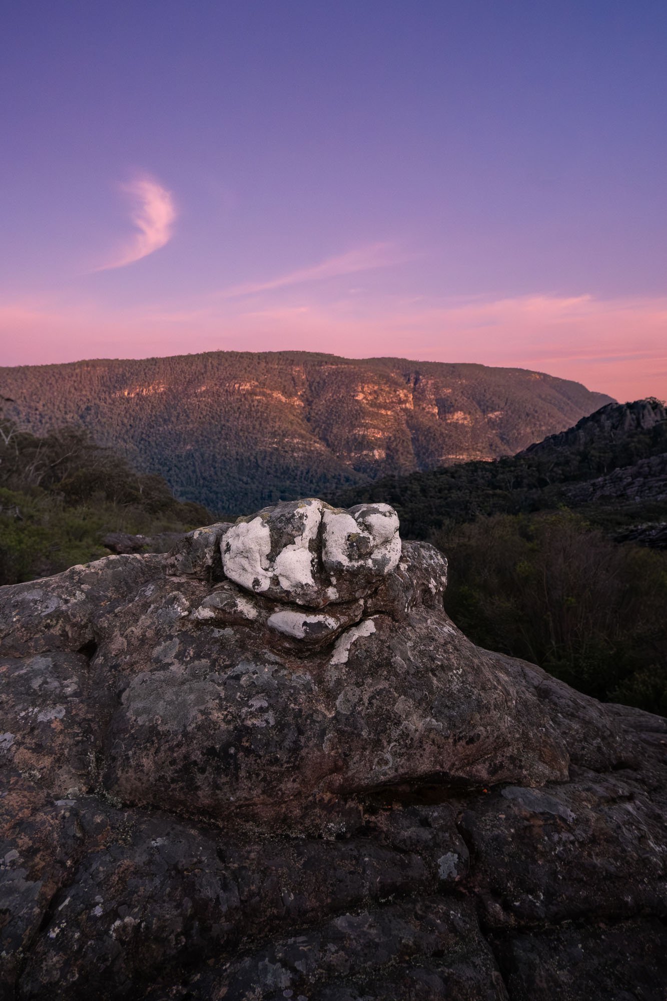 Scenic view of rolling hills with a purple and pink sky at dusk or dawn, rocky foreground with a pile of stones.