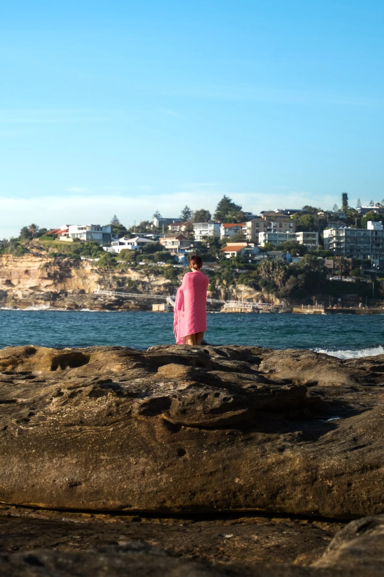 A woman wrapped in a pink blanket standing on rocks by the ocean, overlooking a hillside residential area with houses and trees under a clear blue sky.