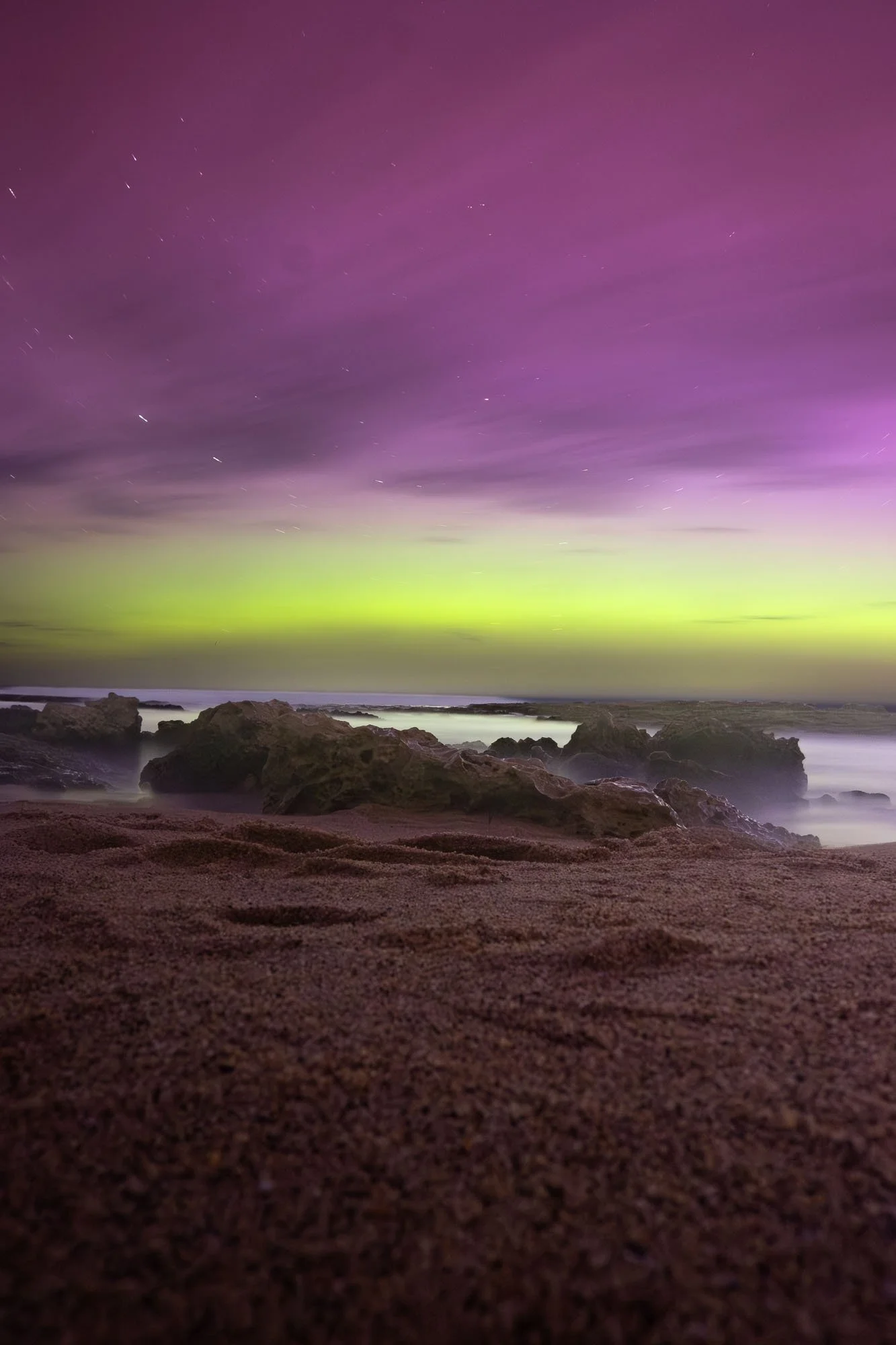 Night sky with purple and green aurora over a rocky beach with sandy foreground.