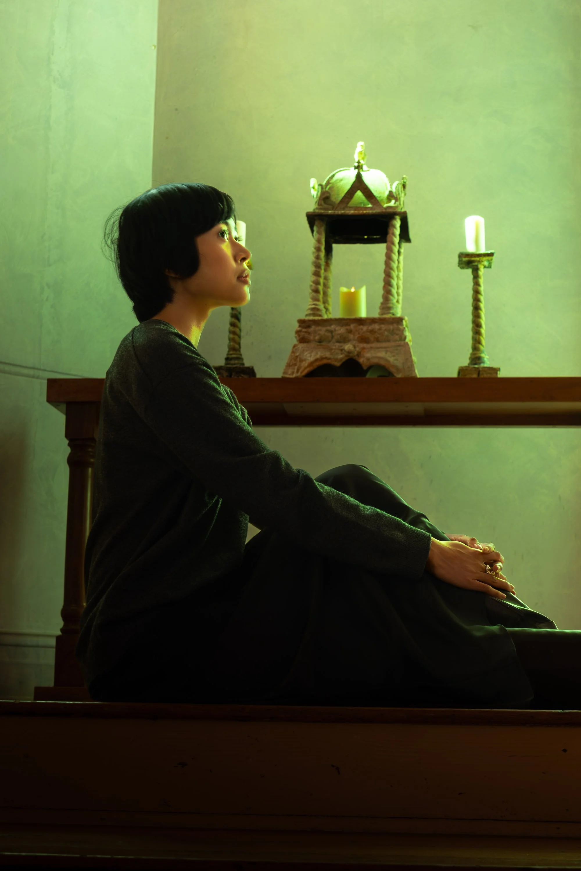 A woman with short black hair sitting on a stair, wearing a dark sweater, with a greenish light illuminating her face, in front of a table with a decorative structure with a crown on top, surrounded by candles.