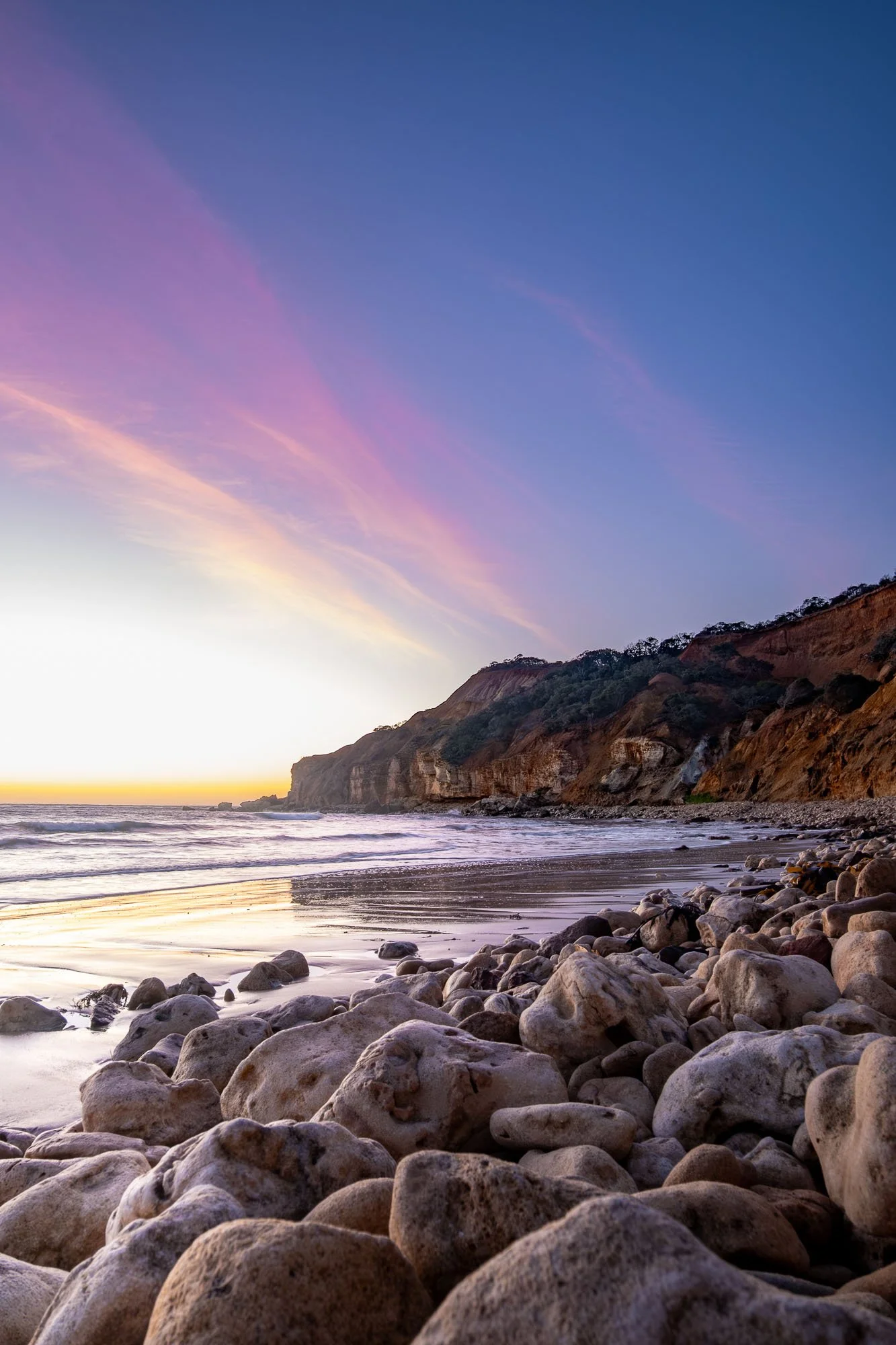 A rocky beach at sunset with the ocean, cliffs covered in greenery, and a colorful sky with pink, purple, and blue hues.