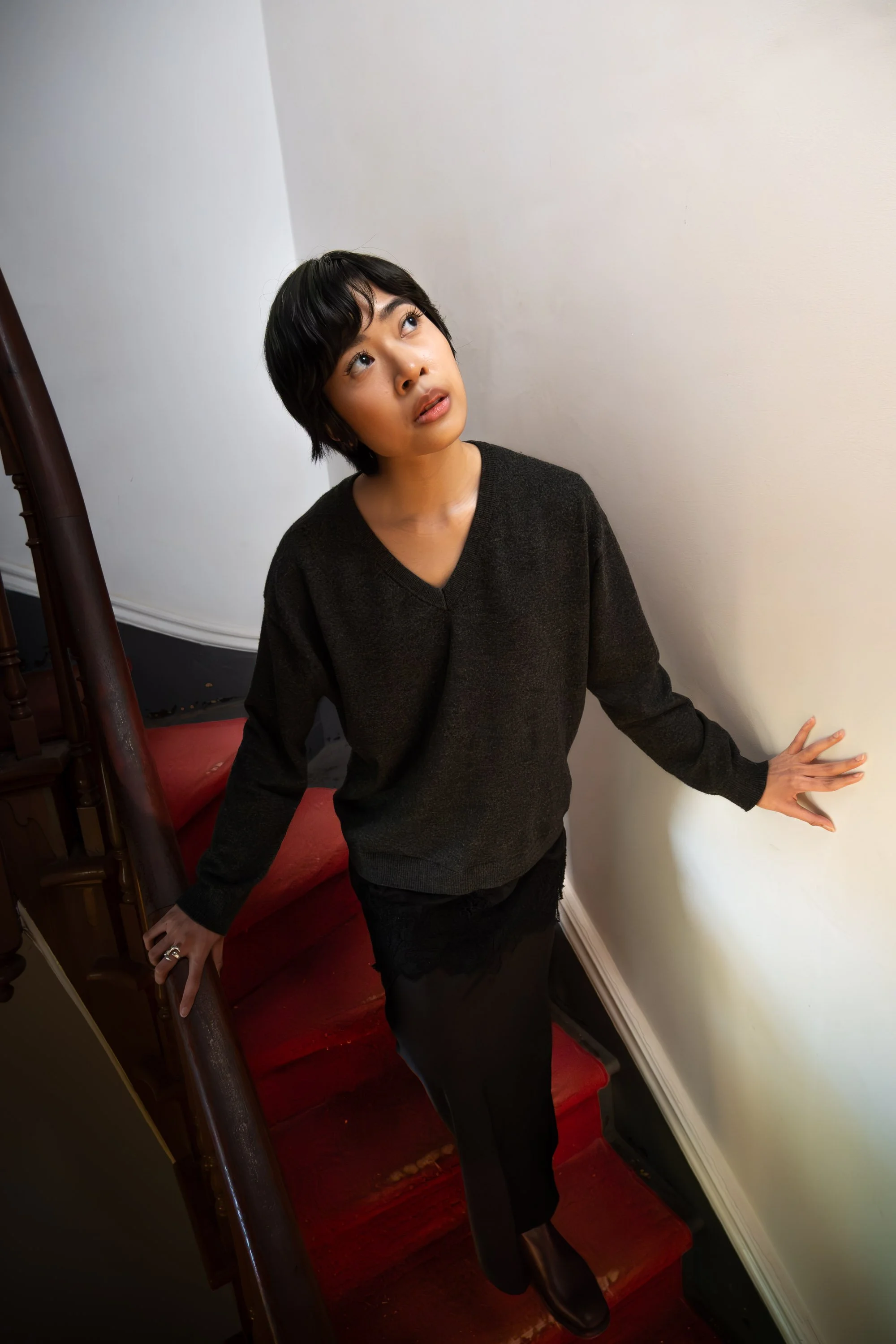 A young woman with short black hair, wearing a black sweater and black skirt, posing on a staircase with red-carpeted stairs and a white wall, looking up and to her left.