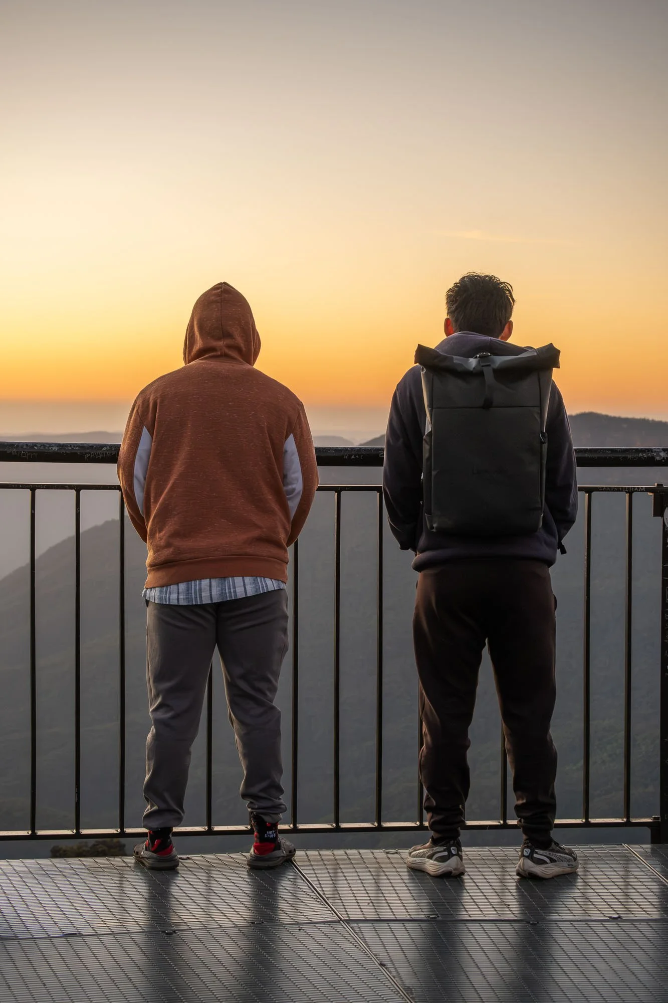 Two people standing on a viewing platform overlooking a mountainous landscape at sunset. One person is wearing a brown hoodie and gray pants, while the other is wearing a backpack, a dark jacket, and black pants.