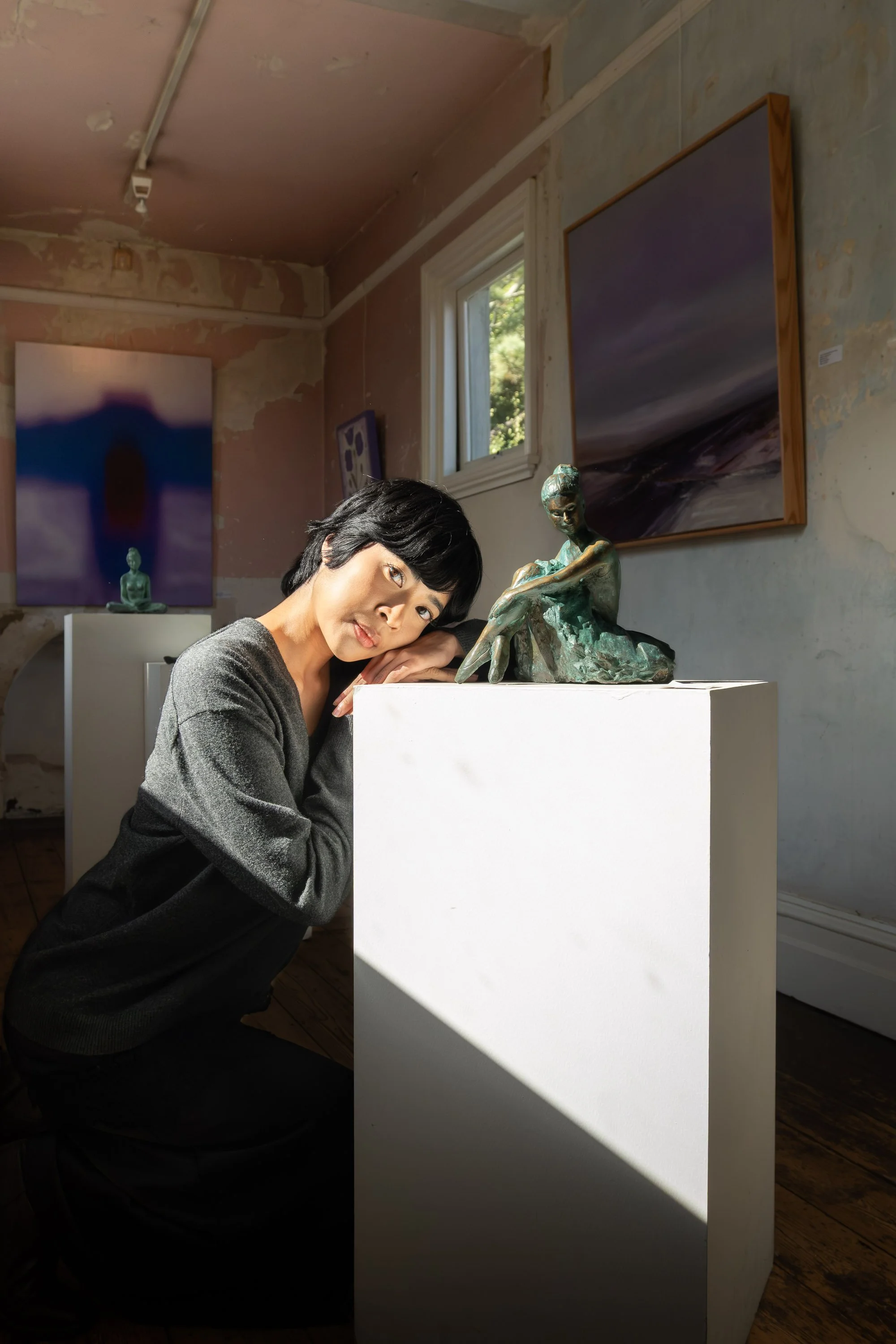 A woman with short black hair and a gray sweater, resting her head on her folded hands on a white pedestal in an art gallery, next to a bronze sculpture of a woman sitting with her knees up to her chest. The gallery has various paintings on the walls