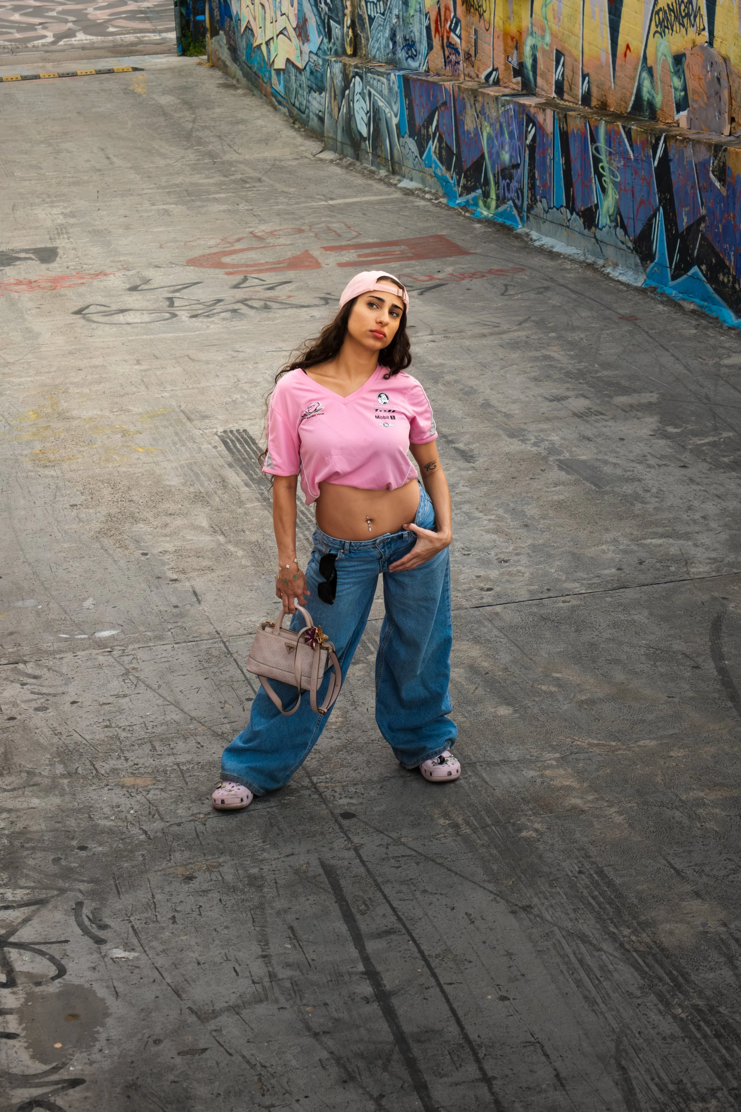 A young woman with dark hair, wearing a pink sports jersey, loose blue jeans, pink Crocs, and a pink cap, stands in an urban alleyway with graffiti-covered walls. She holds a beige handbag in her right hand and has a confident expression.