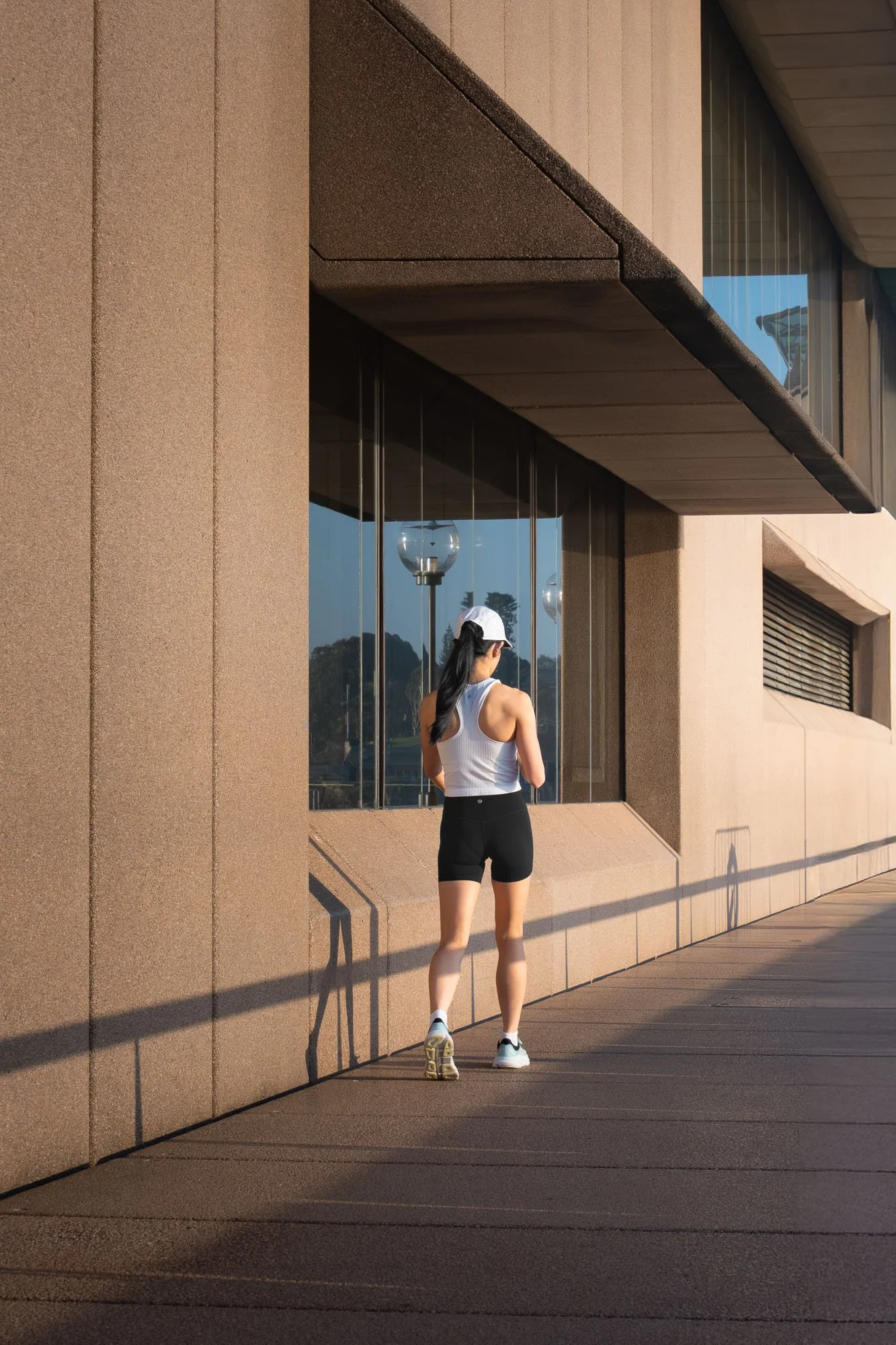A woman in athletic clothing with a white cap is walking along a modern building with large glass windows and textured brown exterior walls during daytime.