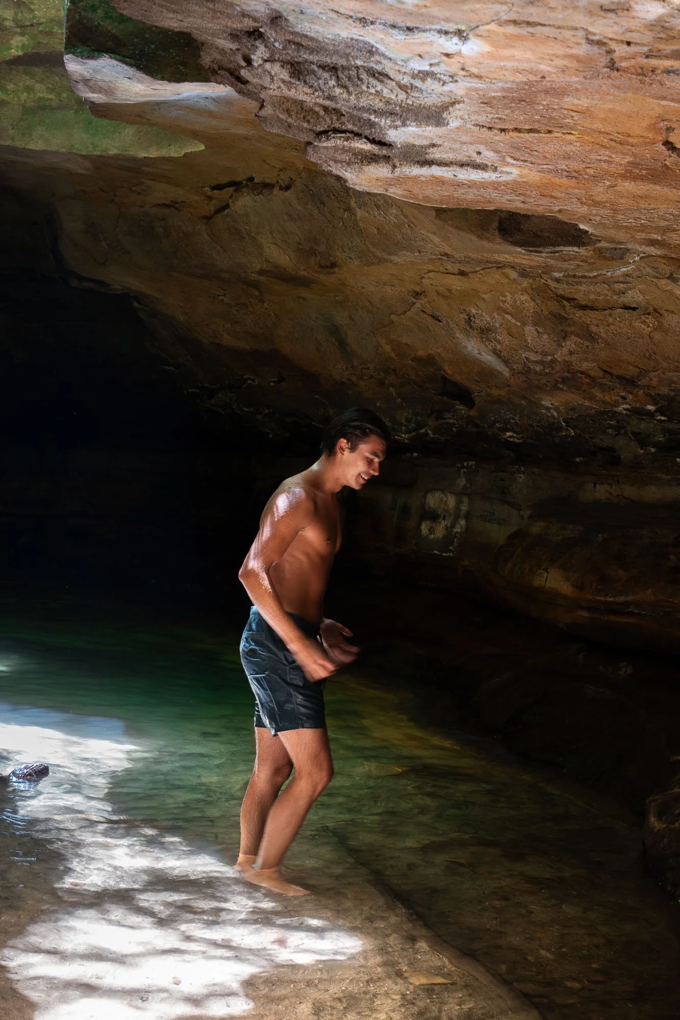 A young man in black shorts stands knee-deep in water inside a rocky cave, smiling and looking downwards, with a curving rock ceiling overhead and sunlight shining through openings.