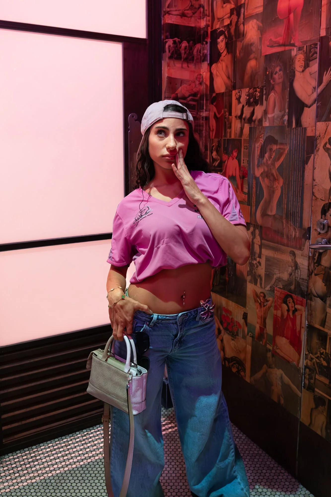 A woman with dark wavy hair wearing a pink cropped T-shirt, blue wide-leg jeans, a beige baseball cap, and jewelry, standing in front of a wall of pin-up style posters, holding a small beige handbag and a water bottle, inside a room with pink lighting and a checkered floor.