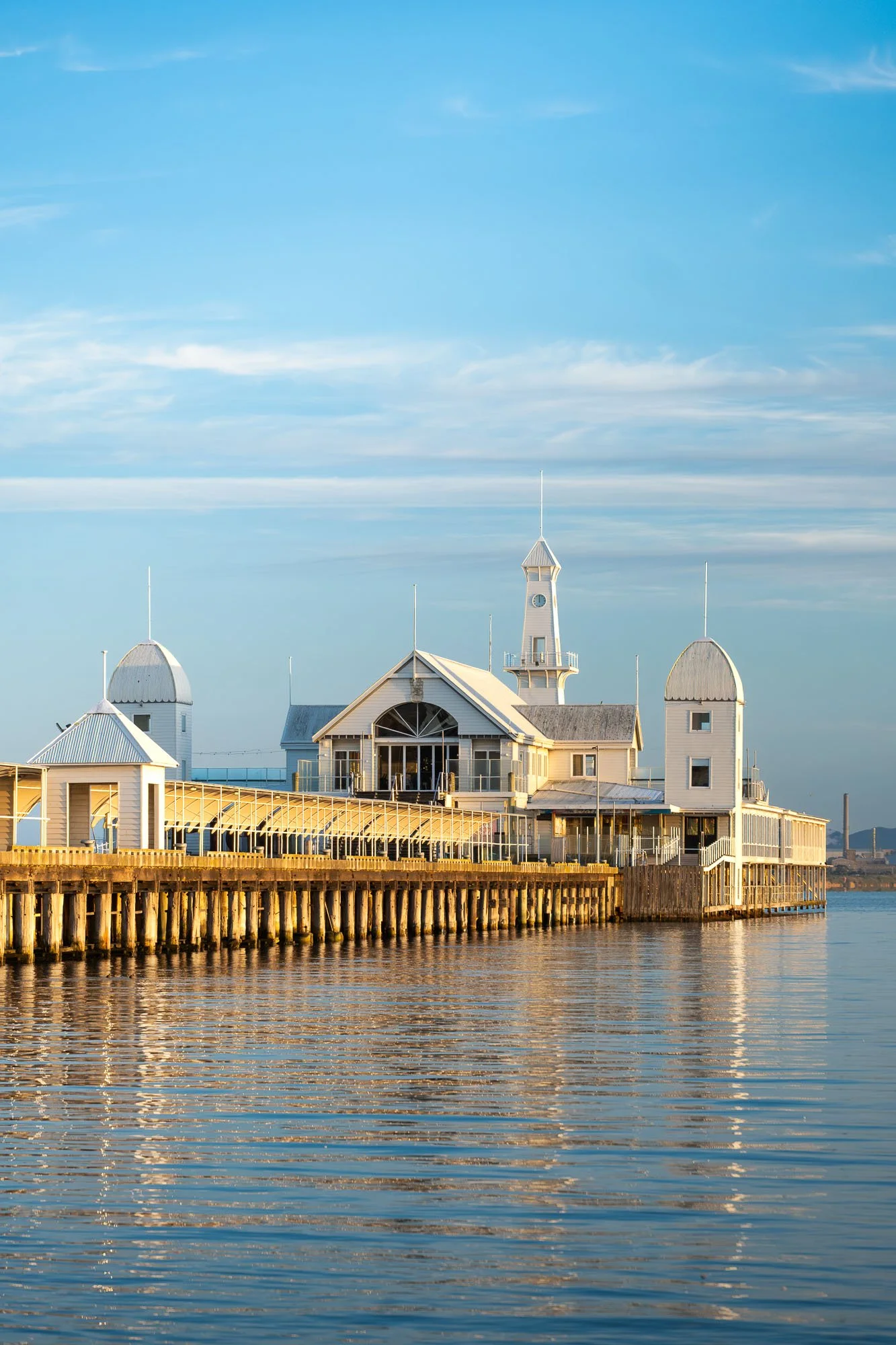A white seaside pier building with a central clock tower, arched windows, and conical domes, extending over water with a reflection and a blue sky with wispy clouds.