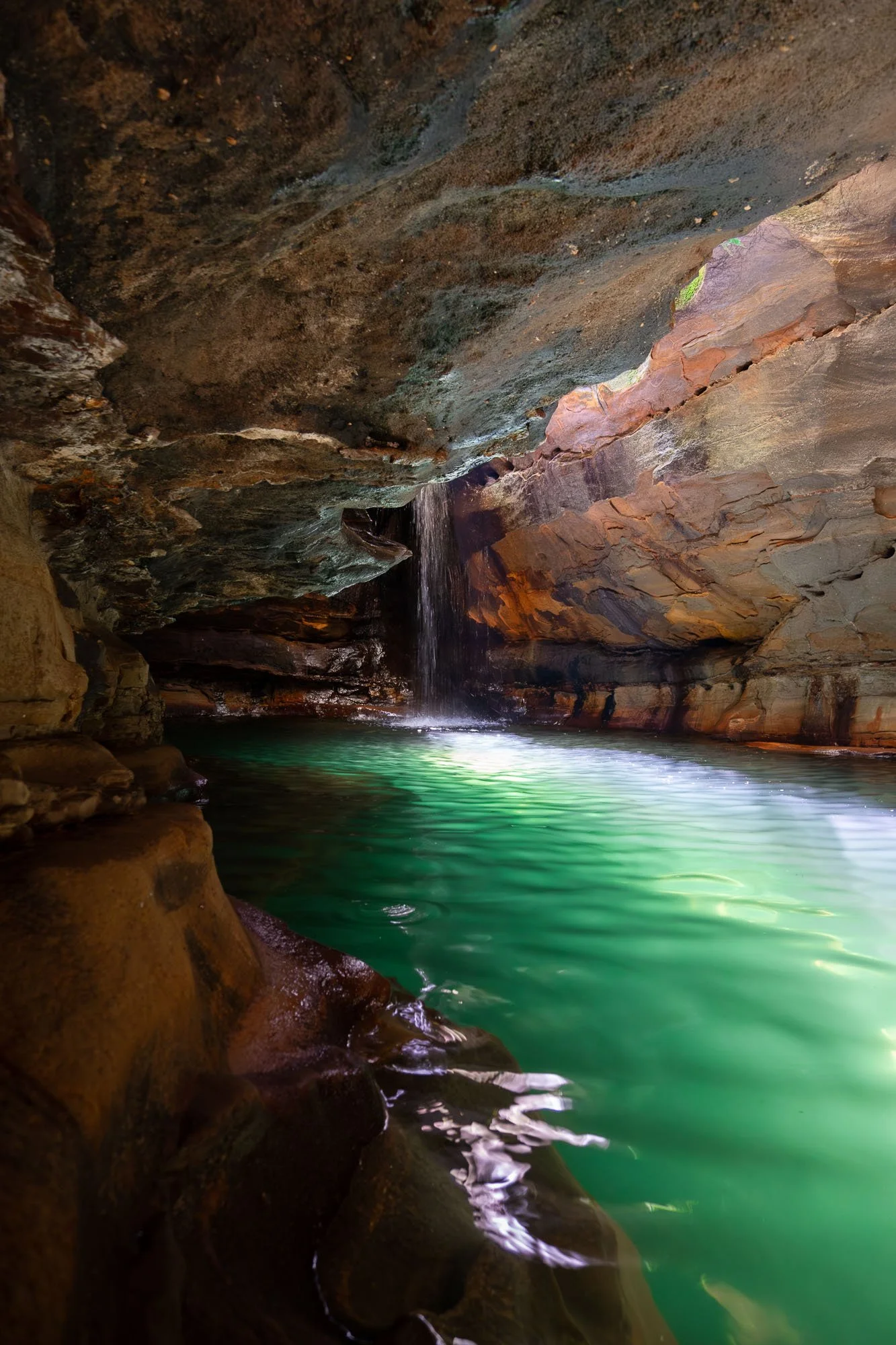 A small waterfall flows into a turquoise pool inside a narrow rocky canyon with reddish-brown rocks overhead.