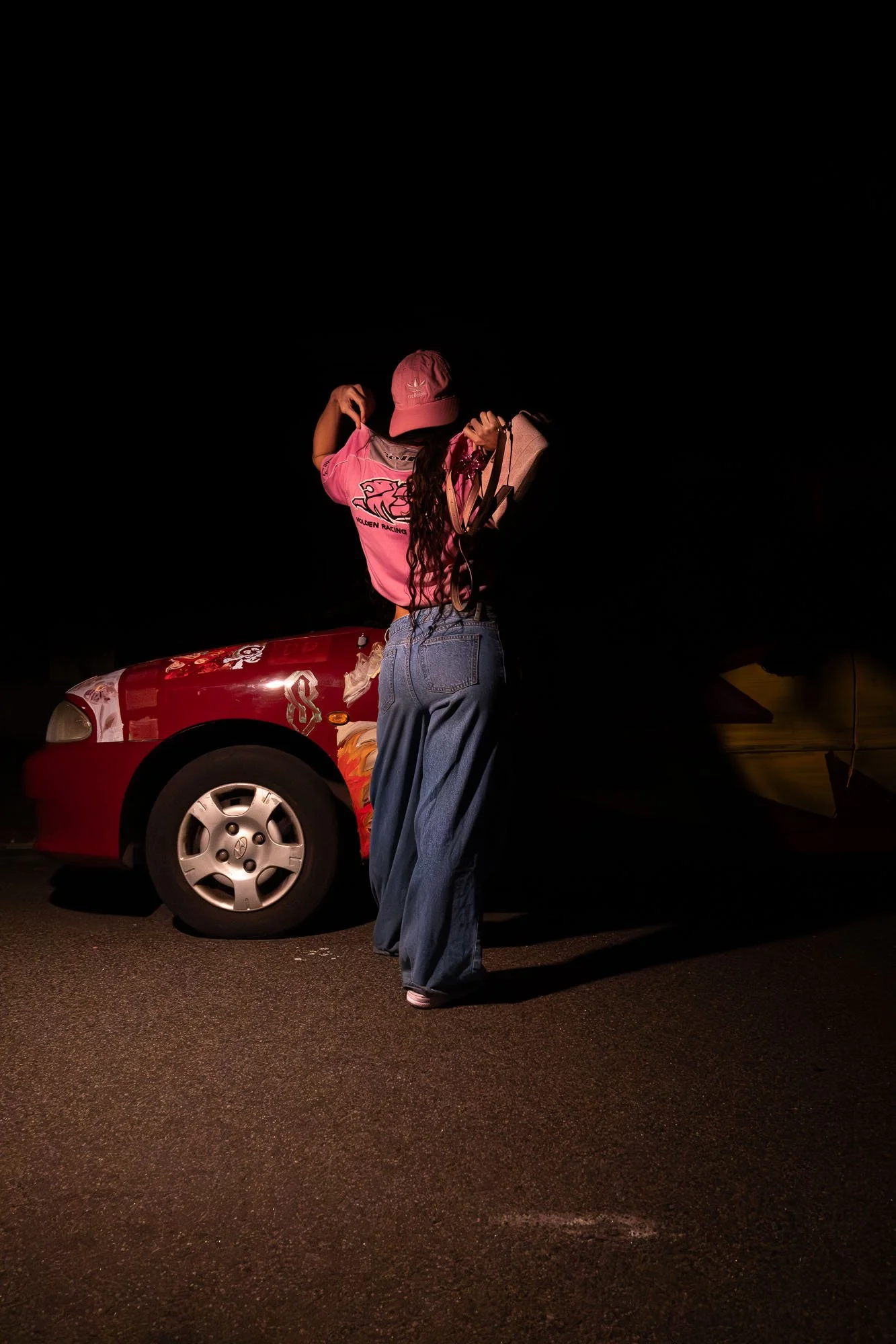 Person standing next to a red race car at night, wearing a pink hat and pink shirt, holding a beige purse.