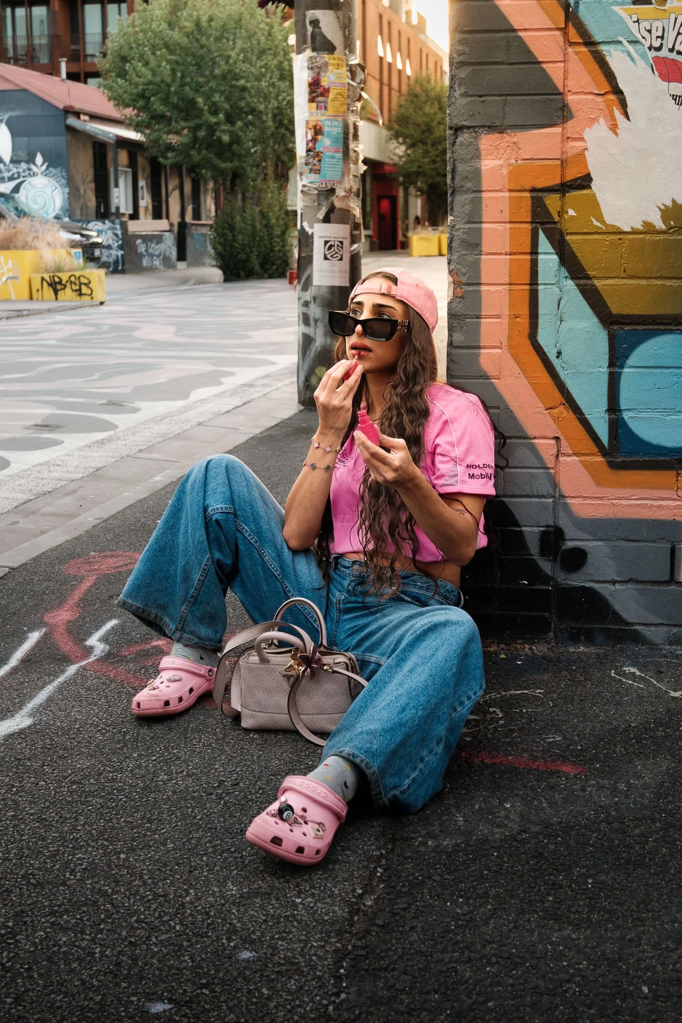 A young woman with long wavy hair, wearing a pink baseball cap backwards, black sunglasses, pink t-shirt, and baggy jeans, is sitting on the sidewalk against a colorful graffiti painted brick wall, holding a lipstick in one hand and applying lip glos