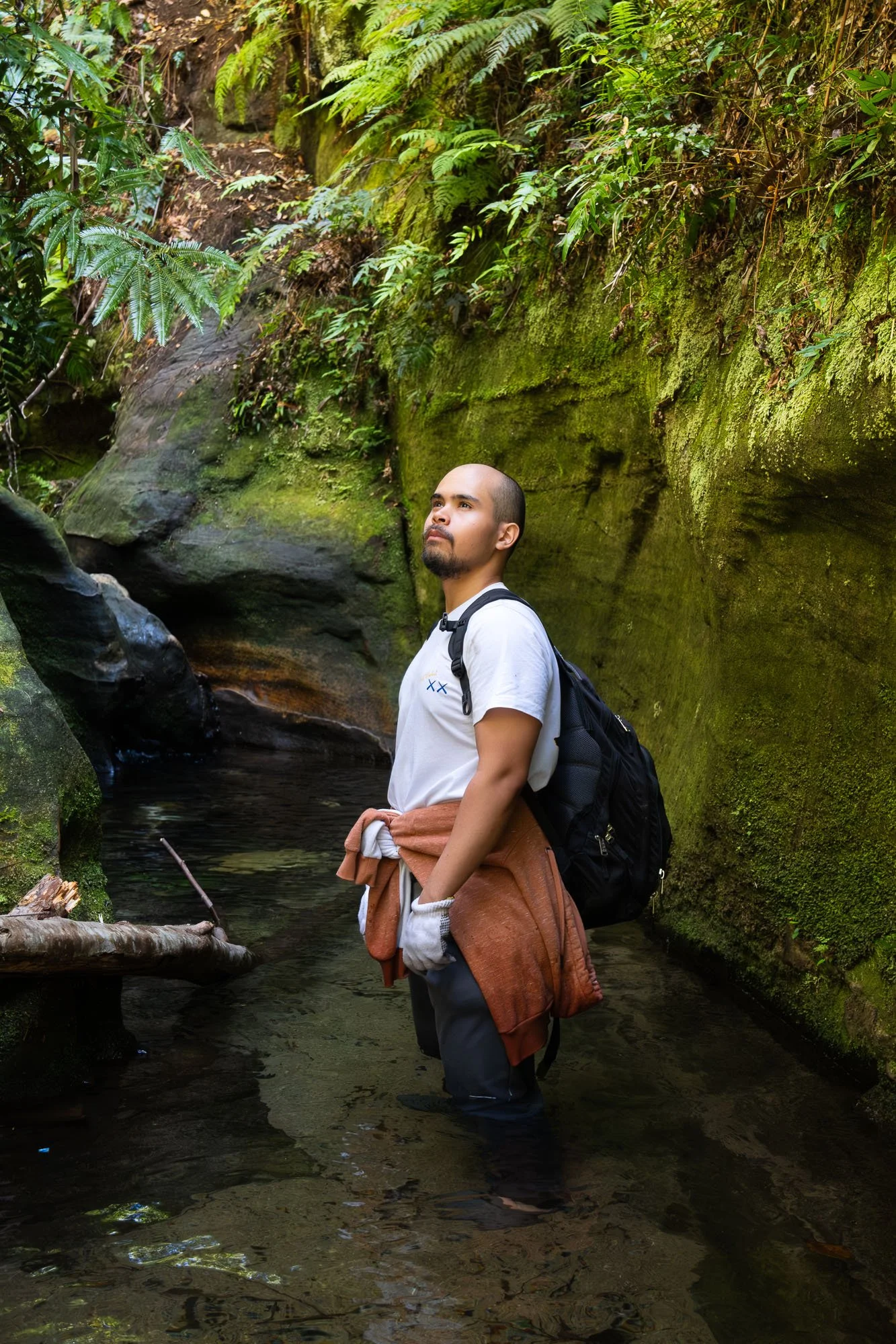 A man wearing a white t-shirt and a backpack stands in a shallow creek, surrounded by green moss-covered rocks and lush foliage, looking up and to the left.