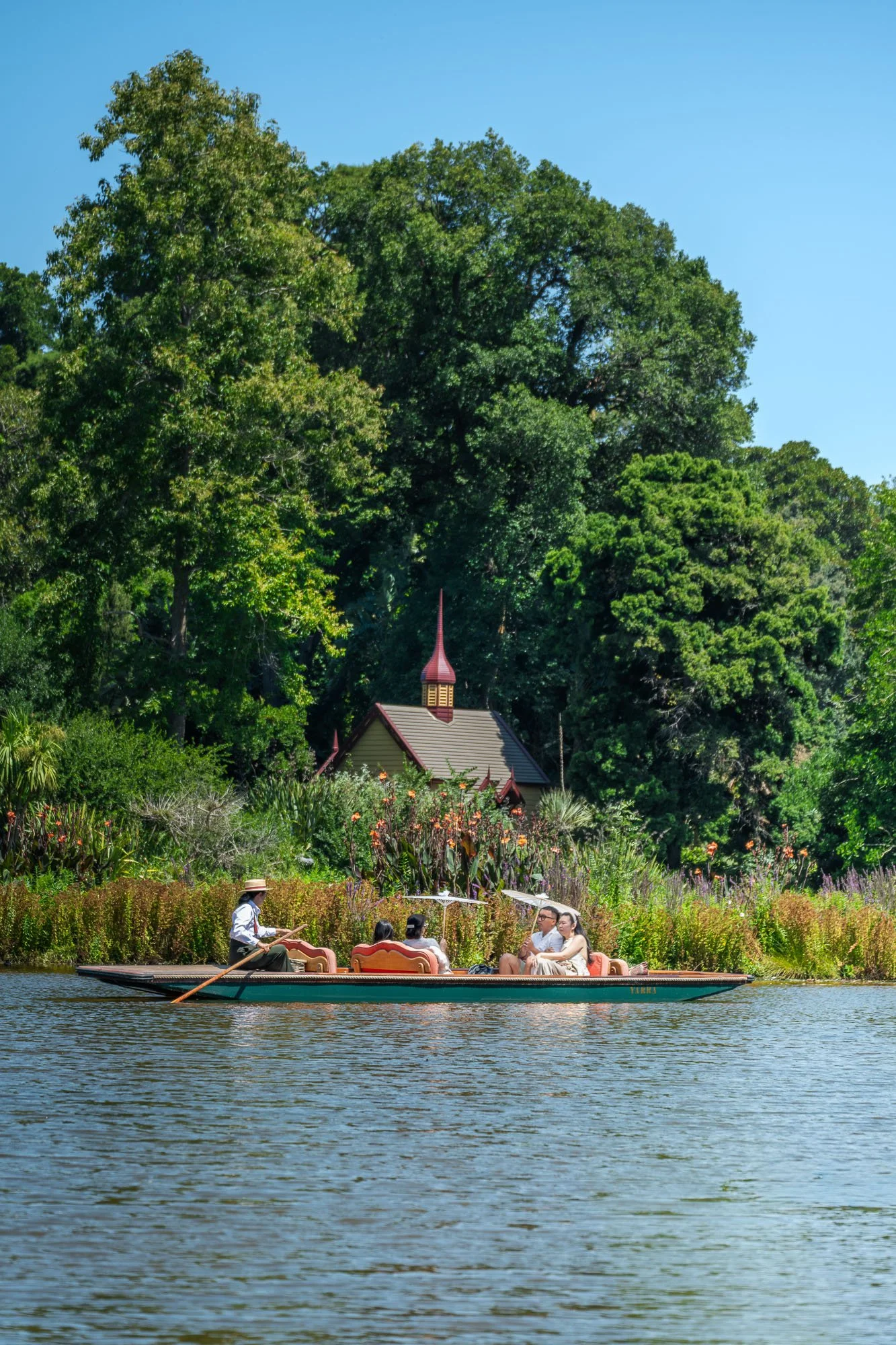 A group of four people enjoying a boat ride on a calm river, with lush green trees, a small church with a red roof and steeple, and a clear blue sky in the background.