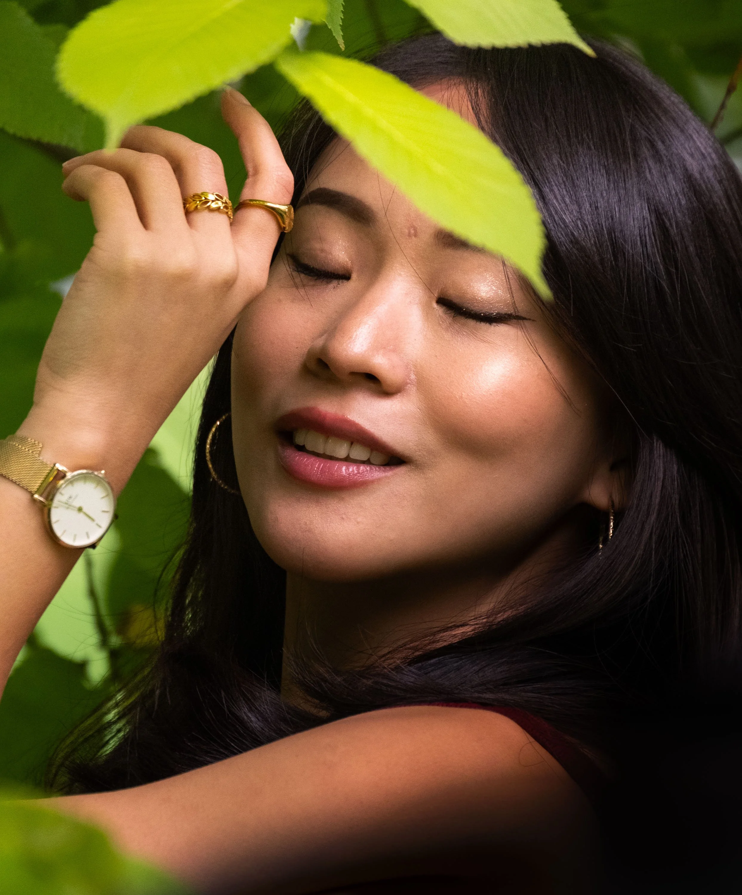 Close-up of a smiling woman with dark hair, closed eyes, wearing gold rings, a gold wristwatch, and hoop earrings, surrounded by green leaves.