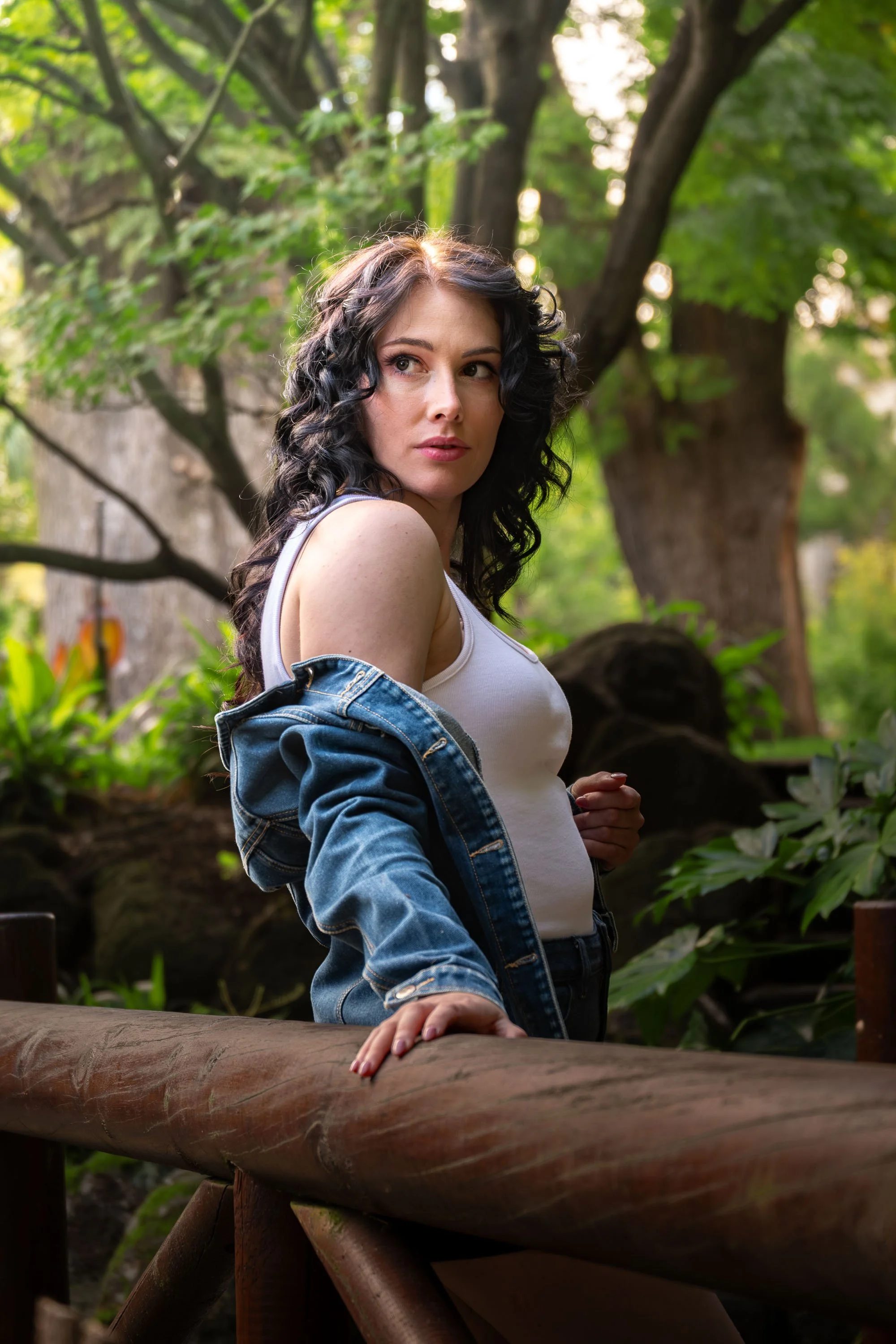 A woman with curly black hair and fair skin is standing outdoors near a wooden railing, looking back over her shoulder. She is wearing a white tank top and a denim jacket, with greenery and trees in the background.