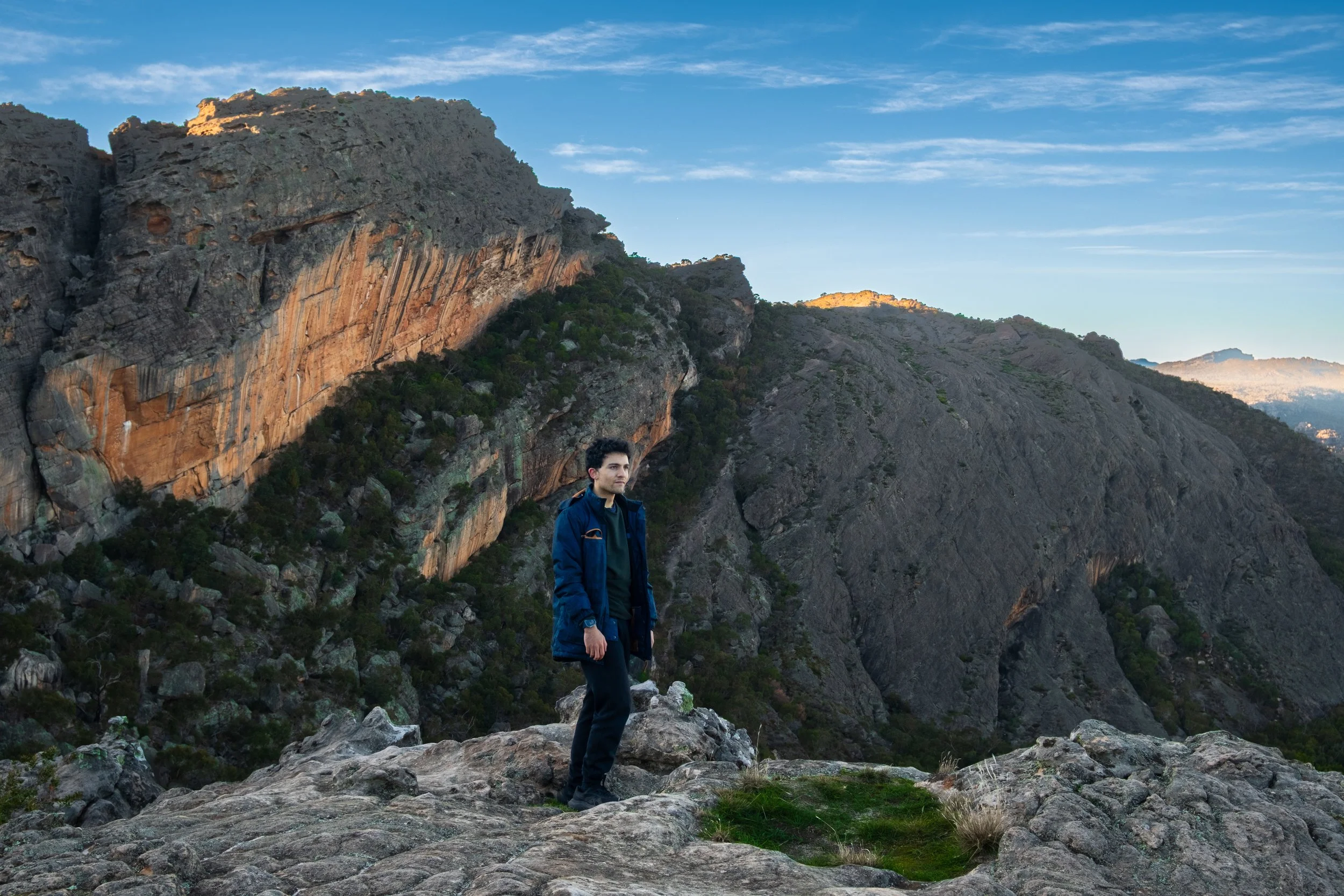 A man standing on rocky terrain in front of rugged mountains with sparse vegetation and a clear blue sky.