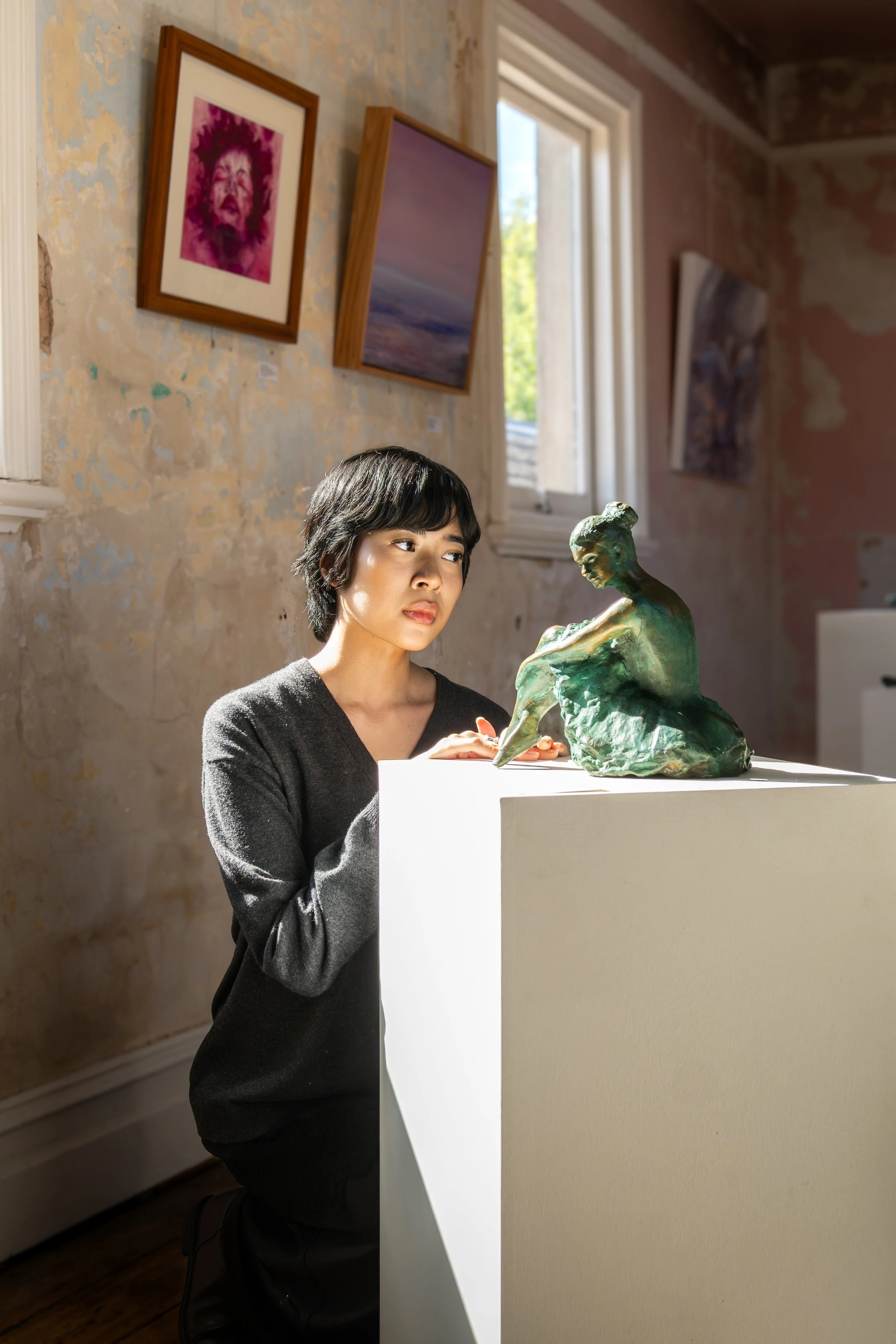A woman with short black hair kneeling beside a sculpture of a woman sitting and holding her knees, displayed on a white pedestal in an art gallery. The gallery has a textured, partially peeling wall with framed artwork and a window letting in natura