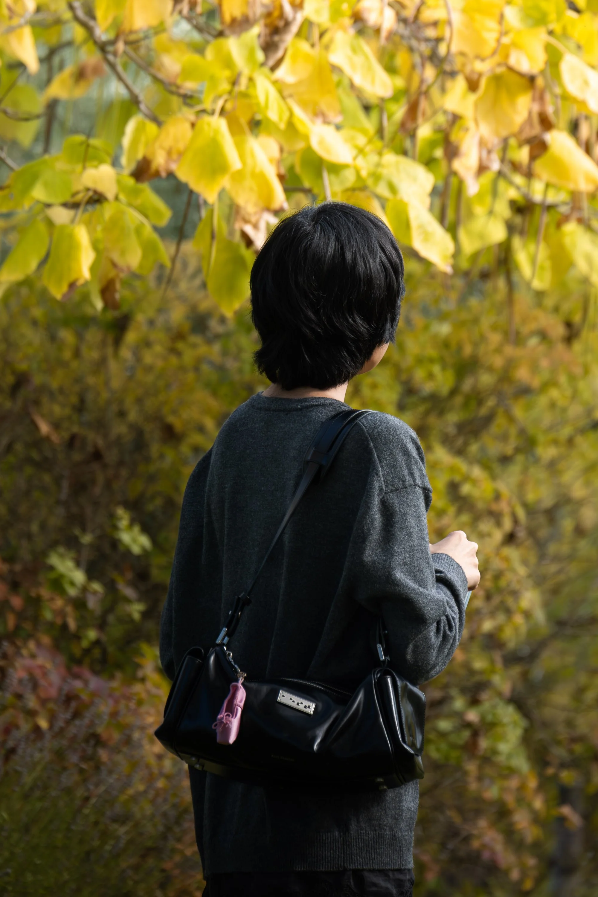Person with black hair wearing a gray hoodie and carrying a black purse with a pink keychain, standing outdoors among yellow and orange autumn leaves.