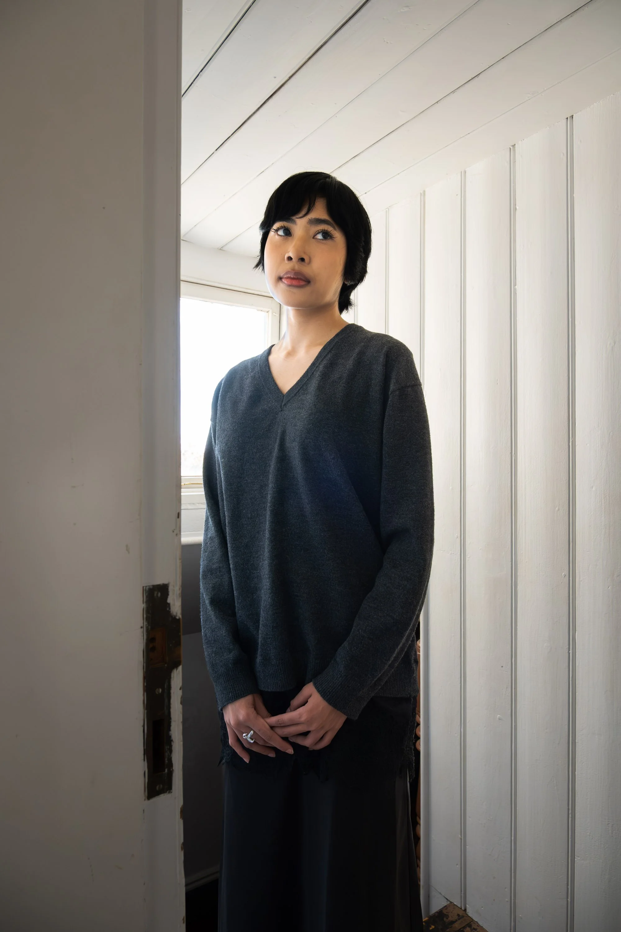 A woman with short black hair wearing a dark gray sweater and black skirt, standing in a white room near a doorway with a window behind her.