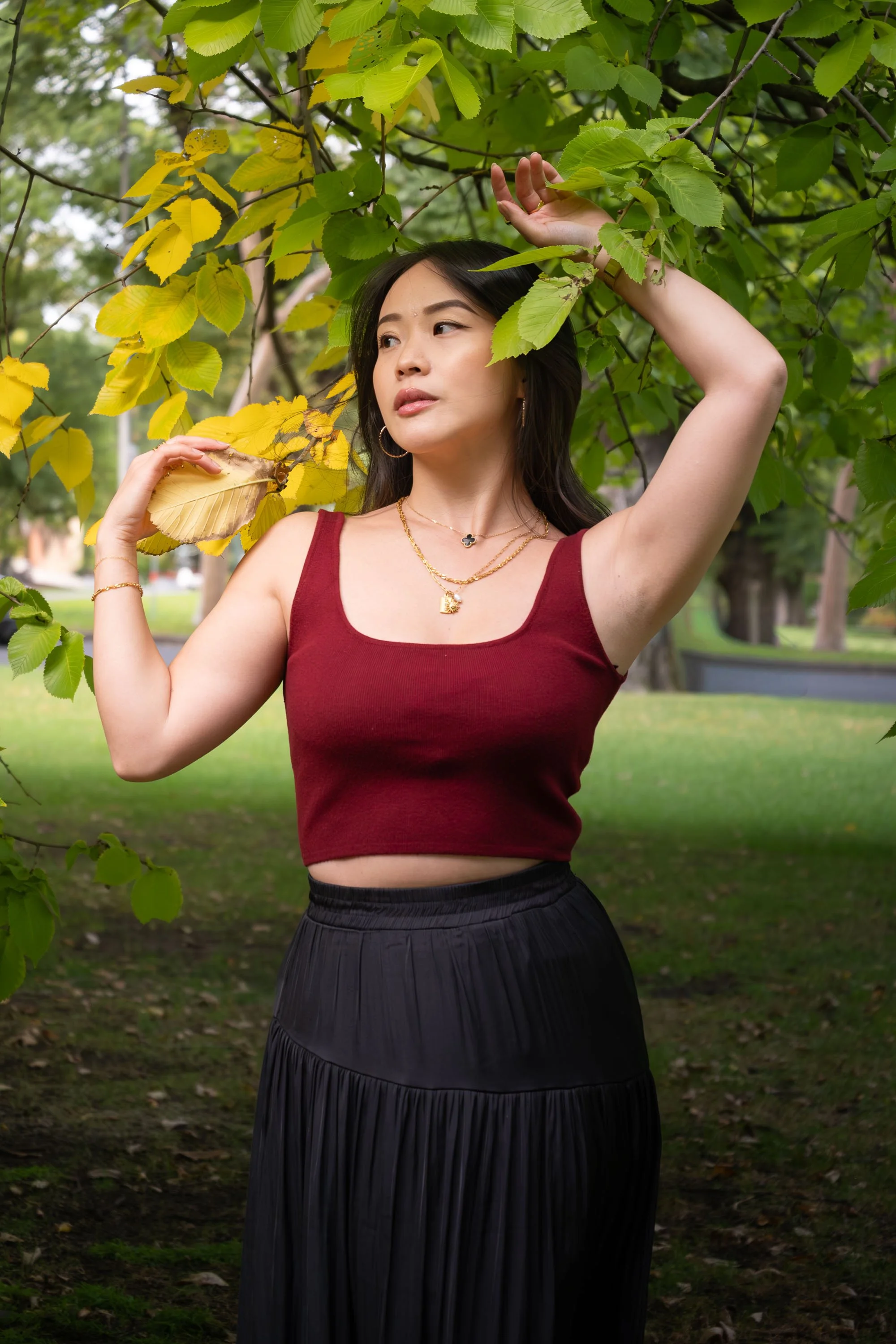 A woman with long black hair wearing a red sleeveless top and black skirt posing outdoors among green trees, holding a leaf near her face and looking to the side.