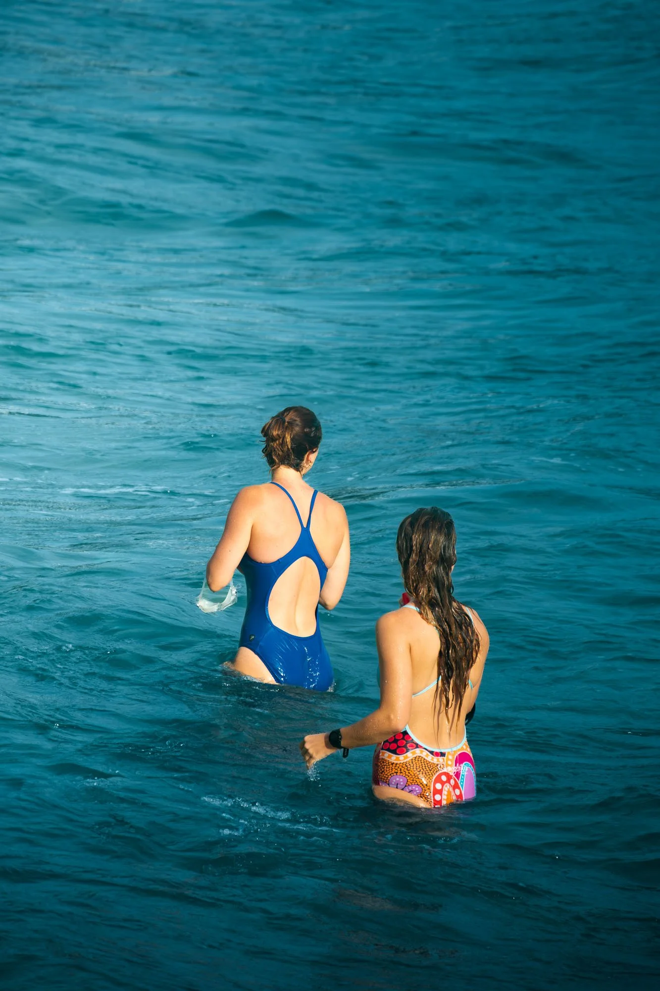 Two women in colorful swimsuits walking into the ocean, with one wearing a blue swimsuit and the other in multicolored swimwear, near the coast with turquoise water.
